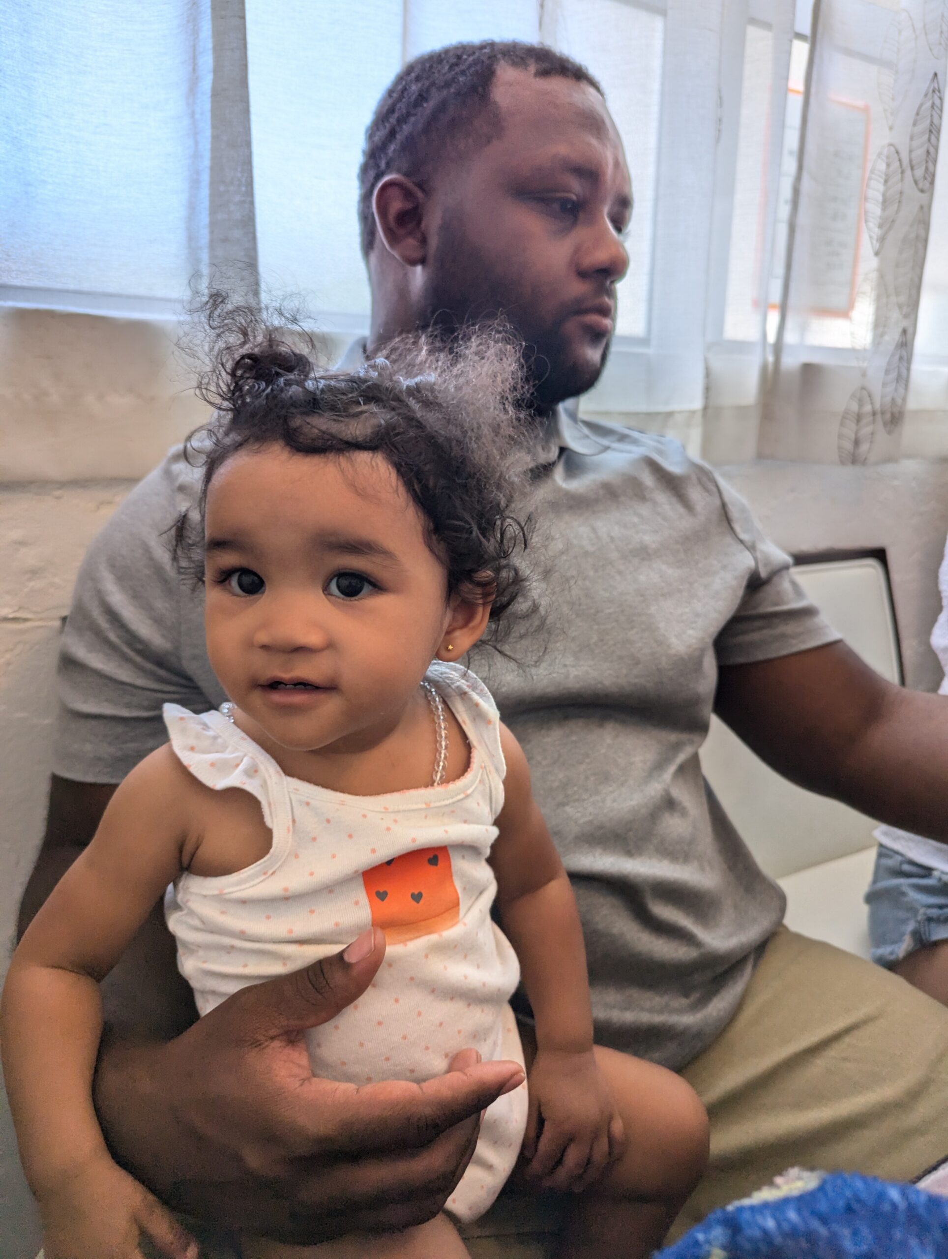 Man wearing a gray shirt sitting indoors, holding a young girl with curly hair on his lap, both looking off to the side near windows.