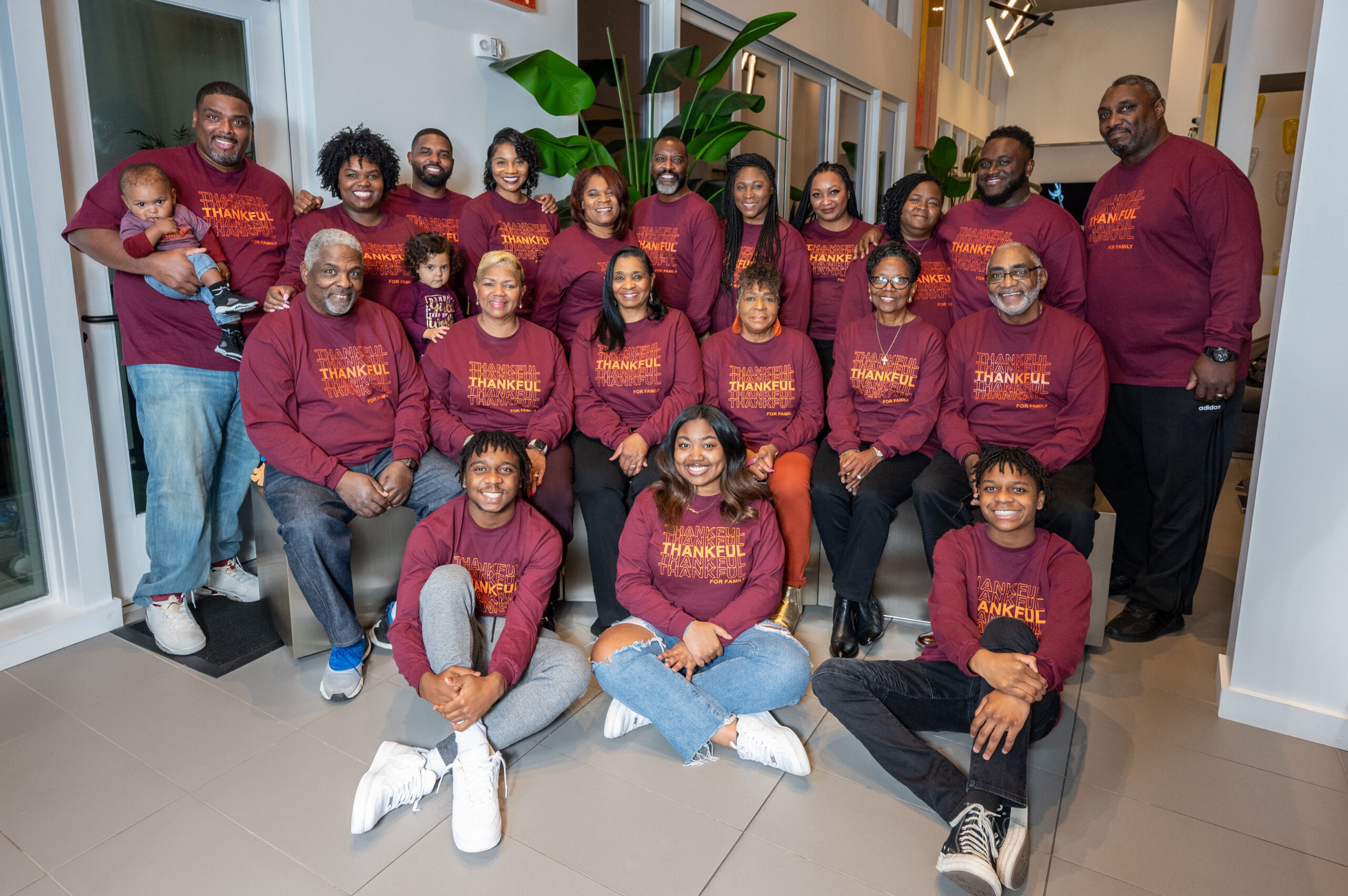 Group of diverse adults and children posing together in a modern office, all wearing maroon long-sleeve shirts that read 'THANKFUL' on the front, smiling for a team photo.