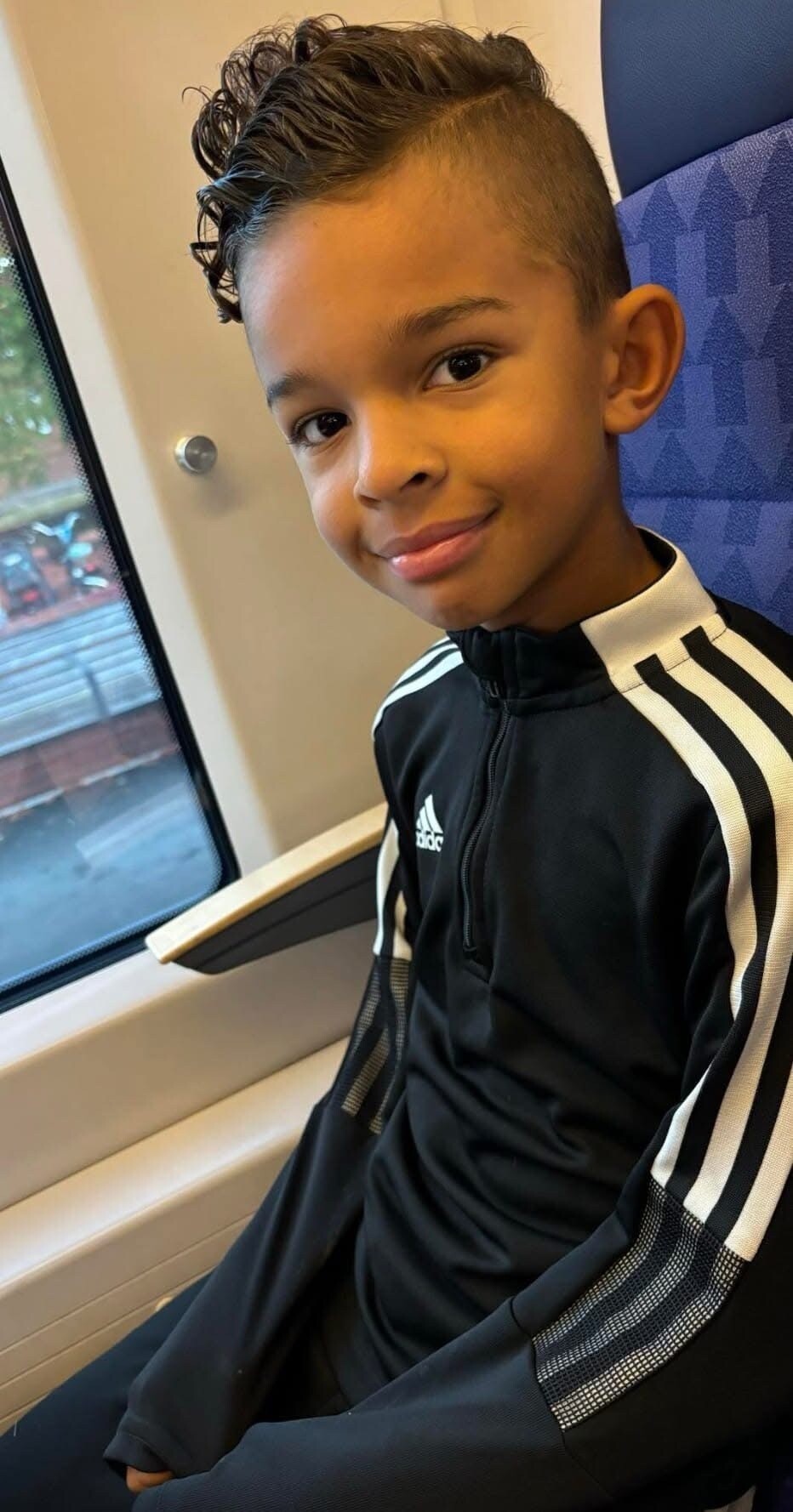 Smiling boy with a mohawk hairstyle sitting on a train in a black Adidas track suit.