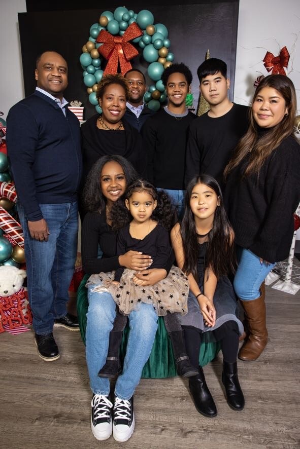A family group portrait posing in front of a Christmas wreath and gifts, with children seated in front and adults standing behind them, all smiling in festive attire.