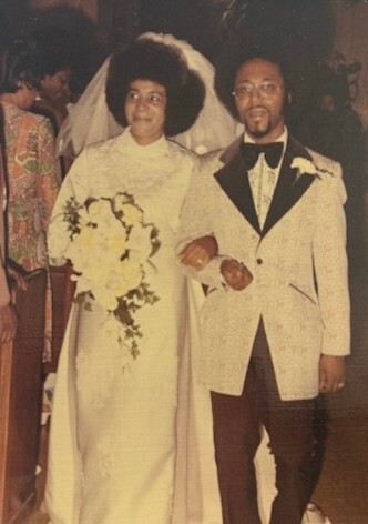 Bride and groom walk arm in arm at their wedding; bride holds a cascading bouquet of white flowers, wearing a white dress and veil, groom in a light-colored tuxedo with a dark bow tie.