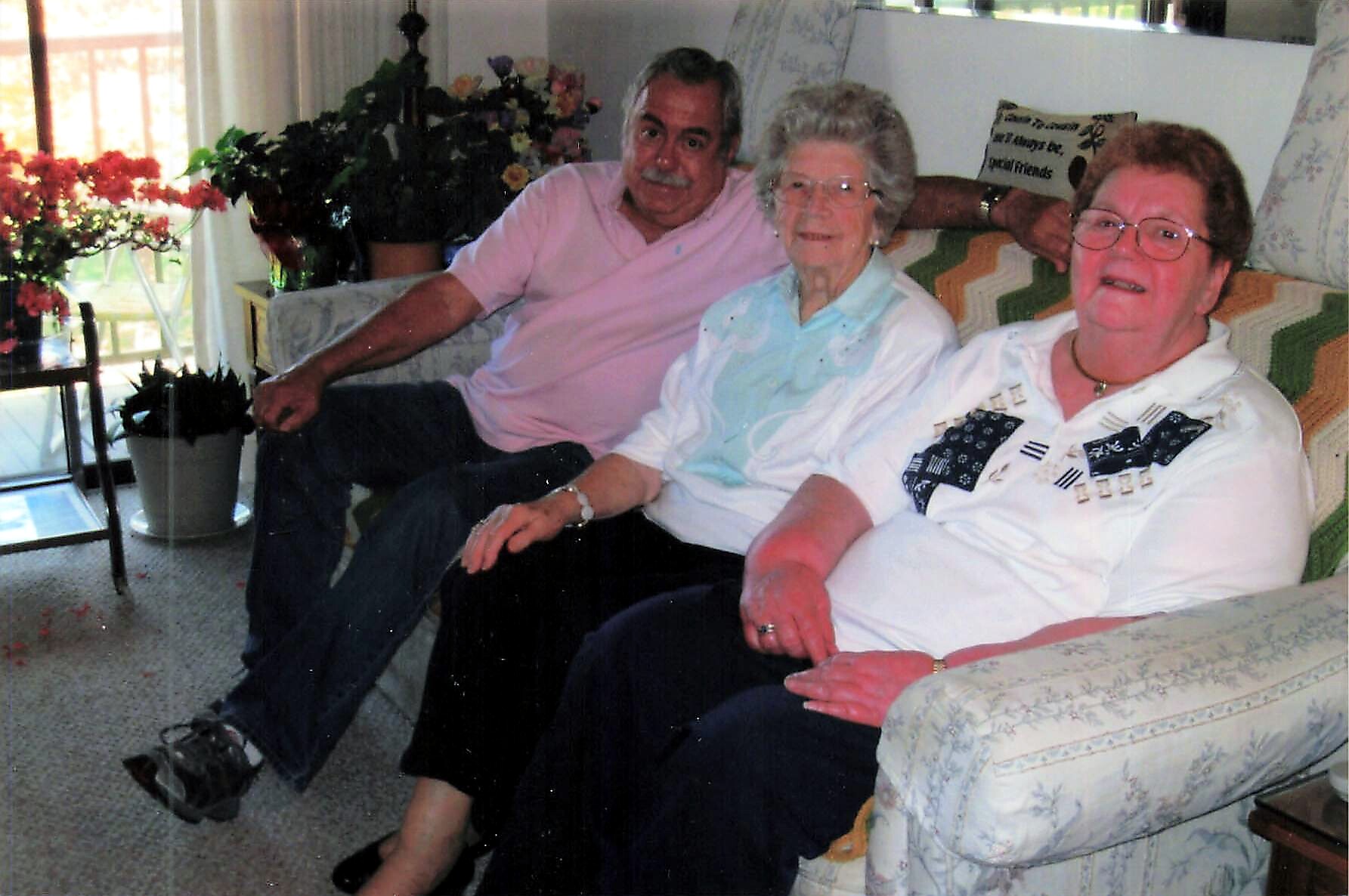 Three elderly adults seated on a patterned sofa in a living room, smiling at the camera with potted plants nearby.