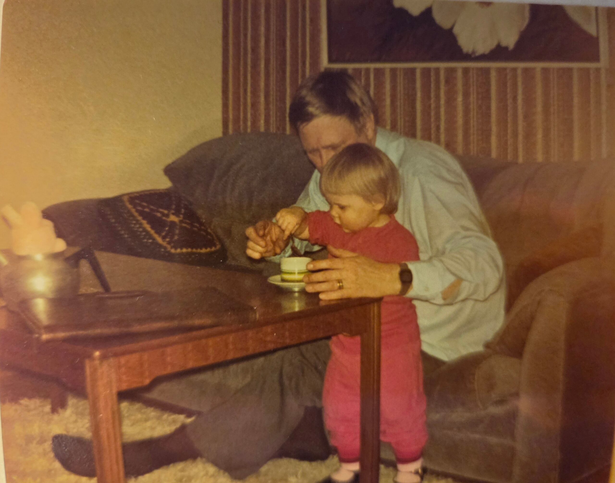 A man in a light blue shirt sits at a table with a small child in red, both focusing on a tiny dessert cup on the table in a retro living room with striped wallpaper.