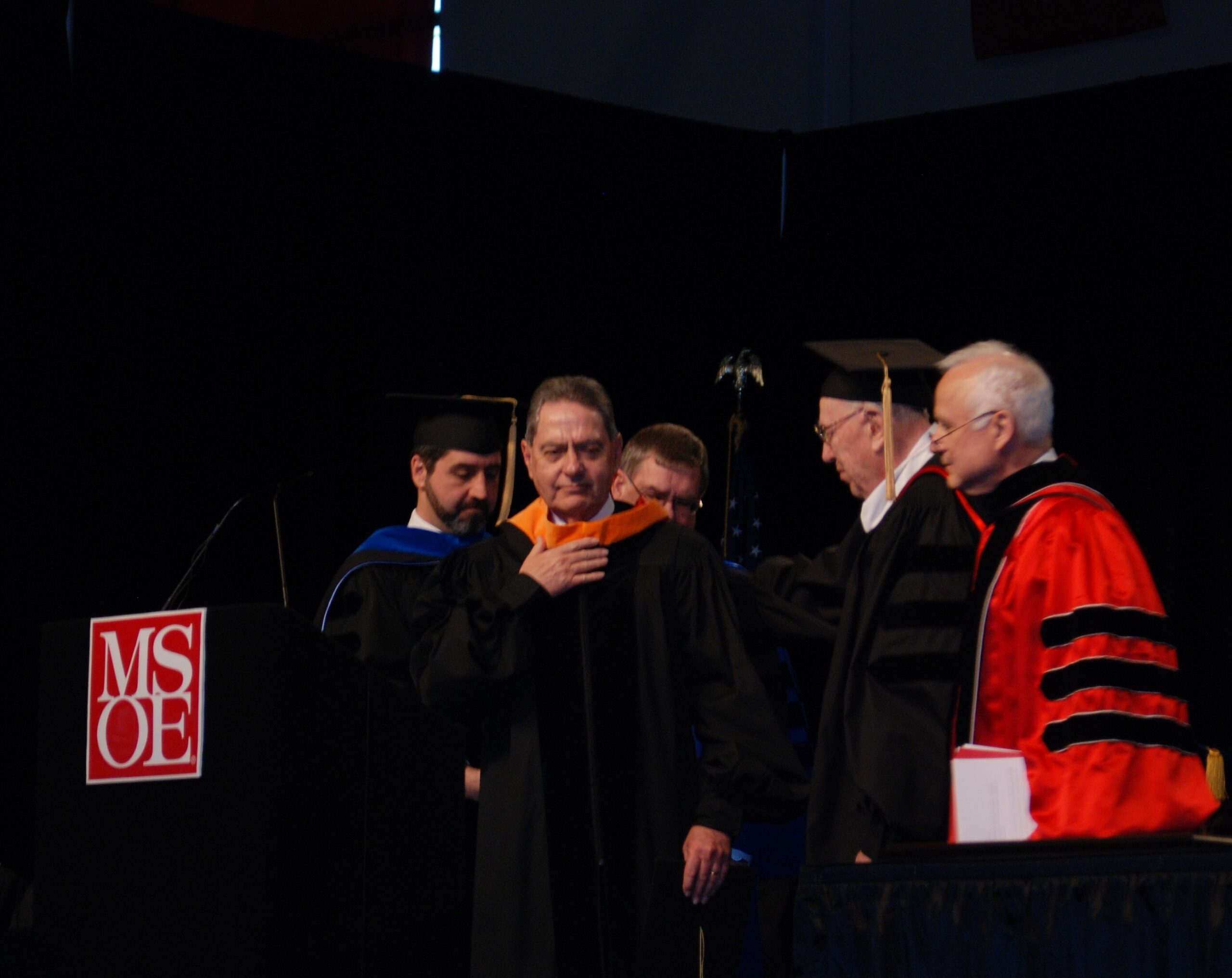 Group of graduates in black gowns on stage during a ceremony, with a red MSOE sign visible in the foreground