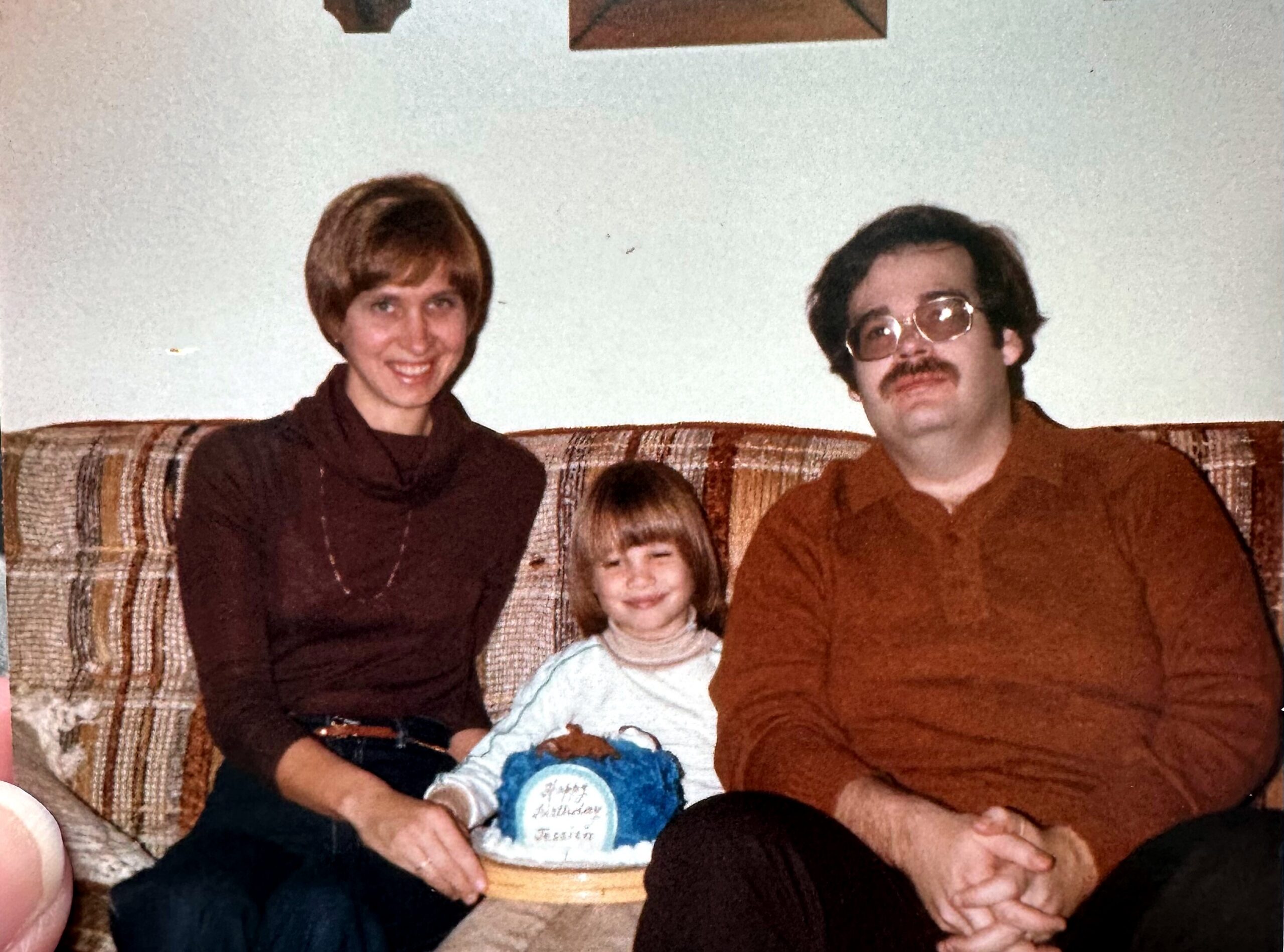 A family of four sits together on a plaid sofa, with a child in the center and a blue birthday cake in front of them.