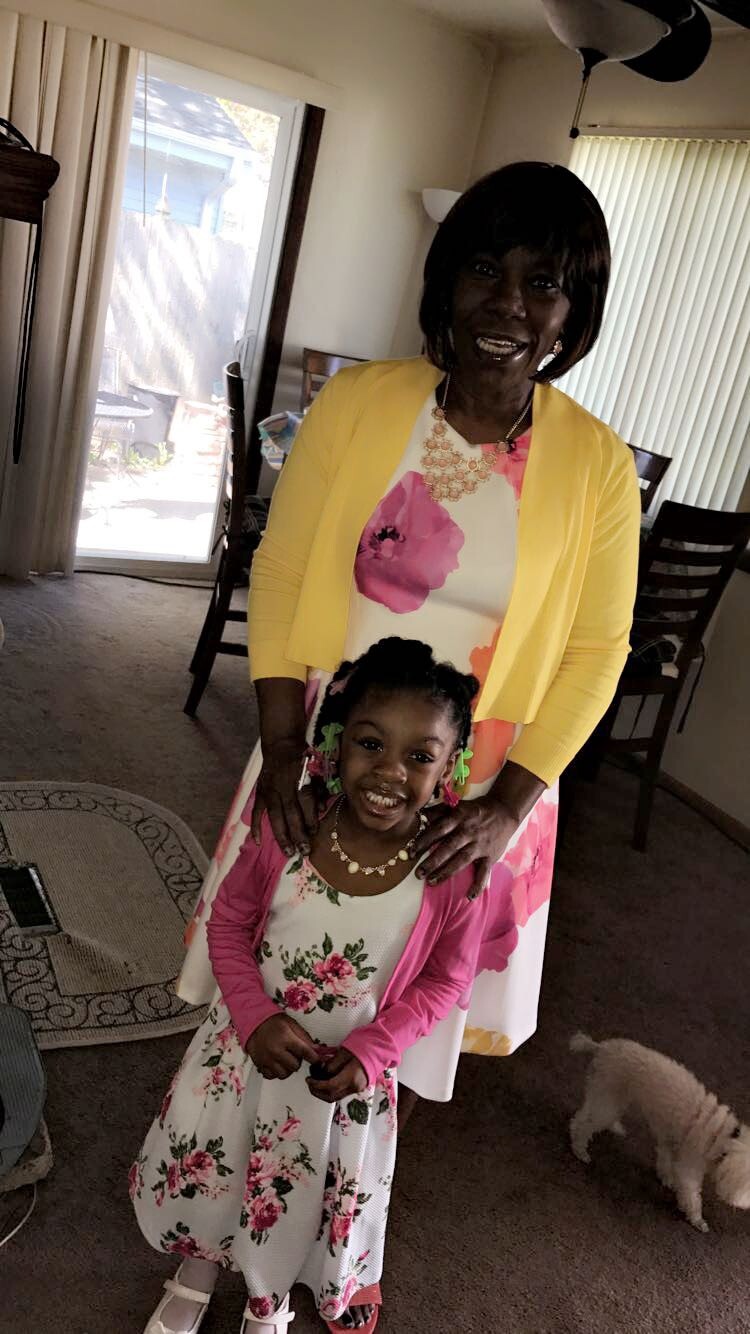 Smiling woman in a yellow cardigan and floral dress stands with a young girl in a matching floral dress in a living room.