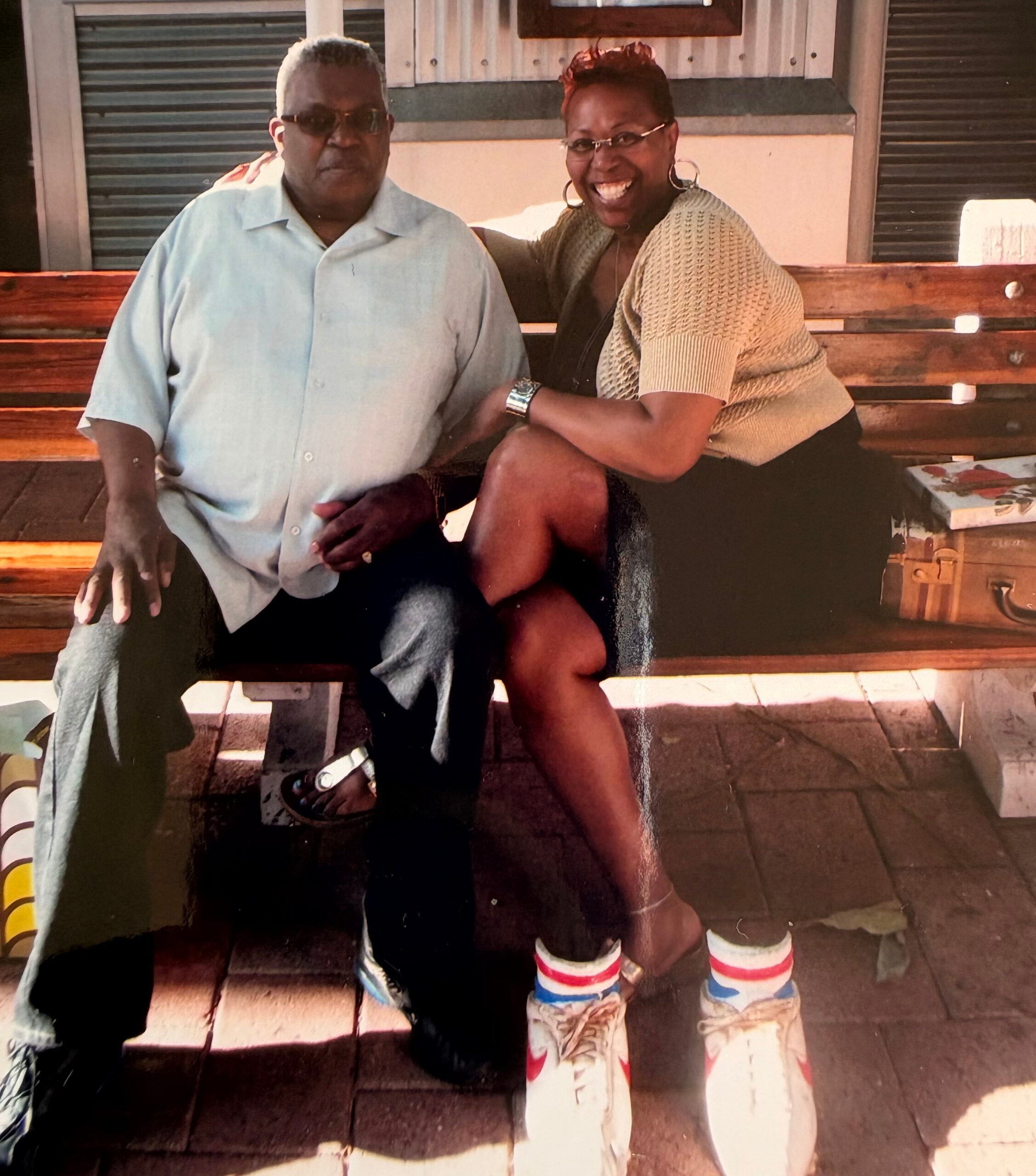 Smiling couple seated on a wooden bench outdoors; man in a light blue shirt and sunglasses, woman in a beige knit cardigan beside him.