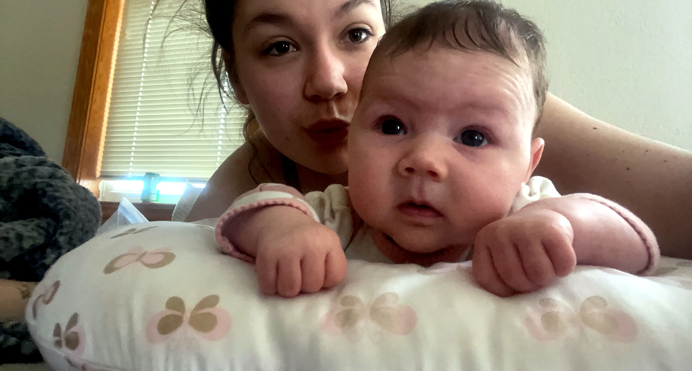 Close-up of a baby in the foreground with a woman (mother) behind, both resting on a bed in a bedroom.