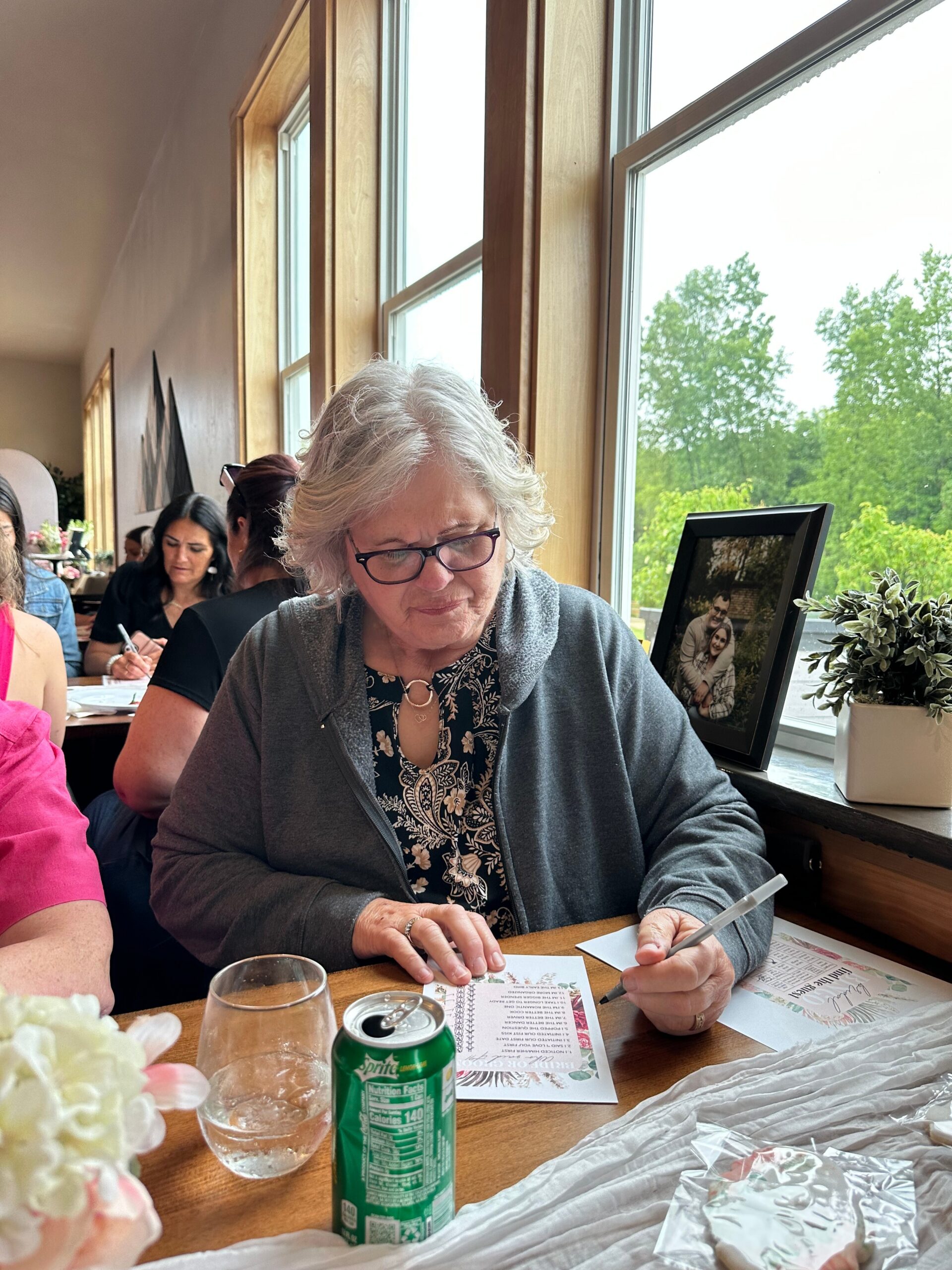 Older woman with glasses signing a paper at a wooden table in a bright cafe, with a Sprite can nearby and a framed photo on the windowsill.