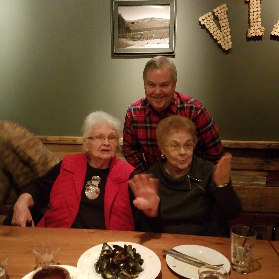 Three adults at a wood-toned restaurant table posing for a photo: two elderly women seated in front, a man in a red plaid shirt standing behind them, with plates of mussels and drinks on the table.