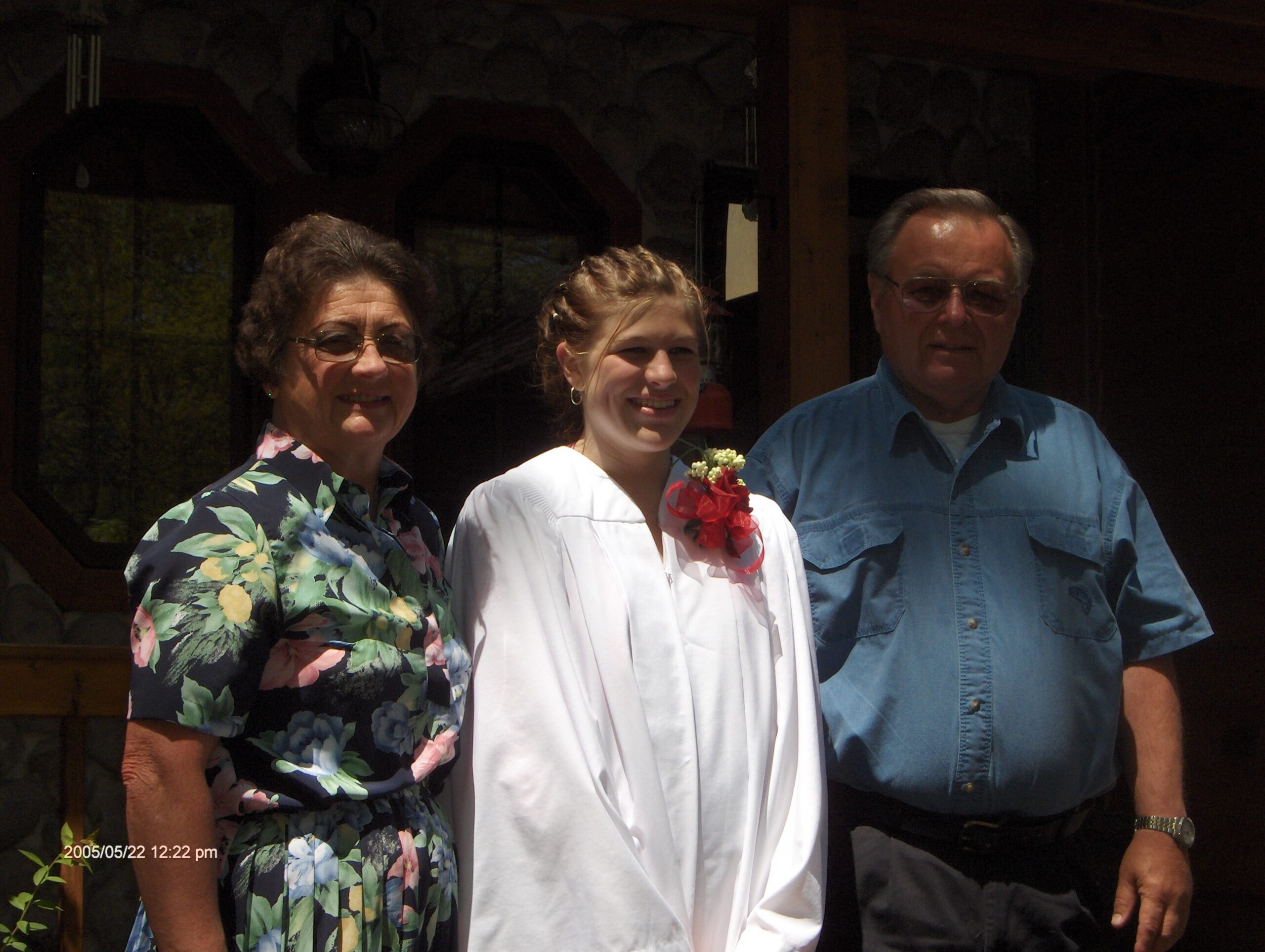 Three people posing outdoors: woman in a floral dress on the left, a girl in white with a red bouquet in the center, and a man in a blue shirt on the right.