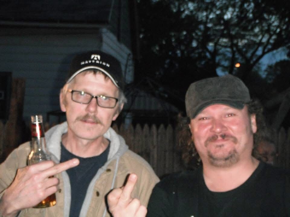 Two men posing outdoors at night; the left man holds a beer bottle and shows a two-finger gesture.
