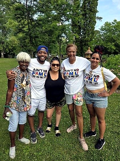 Five people of diverse ages standing together in a sunny park, arms around each other and smiling; three are wearing matching white T-shirts with the slogan 'Ain't No Family Like the One We've Got,' with others in casual outfits.