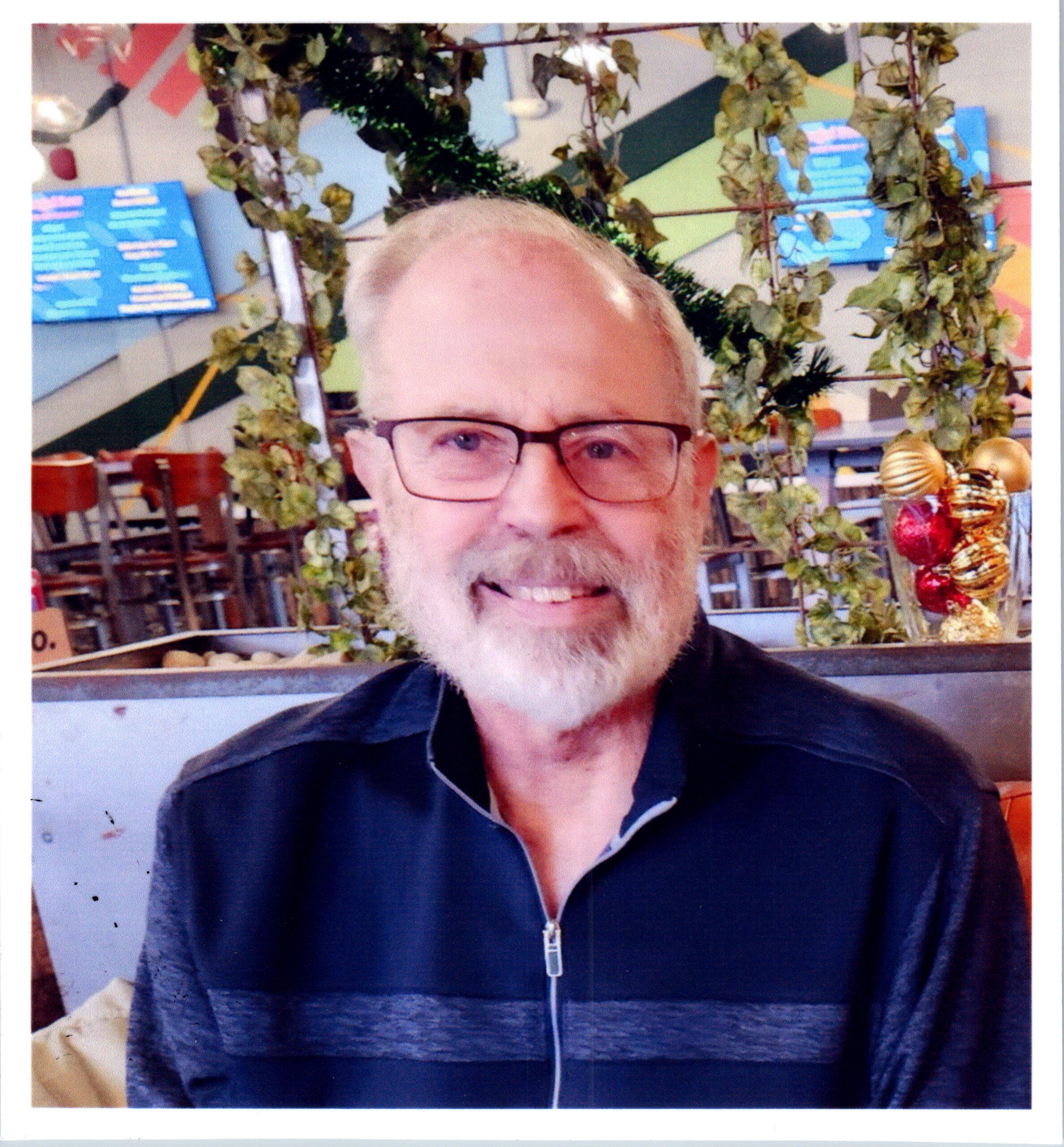 Smiling man with glasses and a white beard, wearing a dark zip-up shirt, indoors with greenery and shelves in the background.