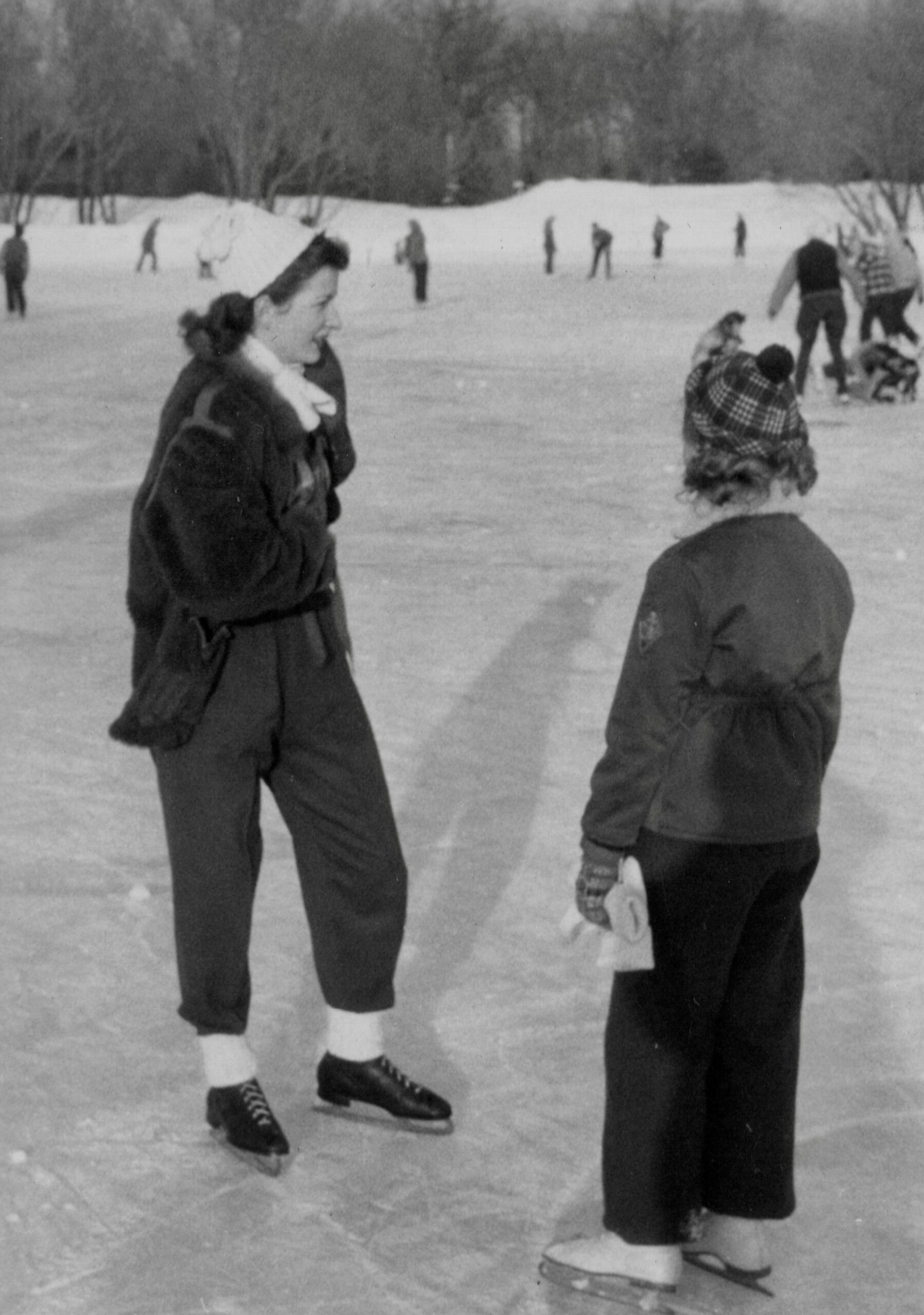 Two people ice skating on a snowy outdoor rink; a woman in a dark coat and scarf stands left while another person with a plaid blanket stands right in the foreground, with more skaters in the background.