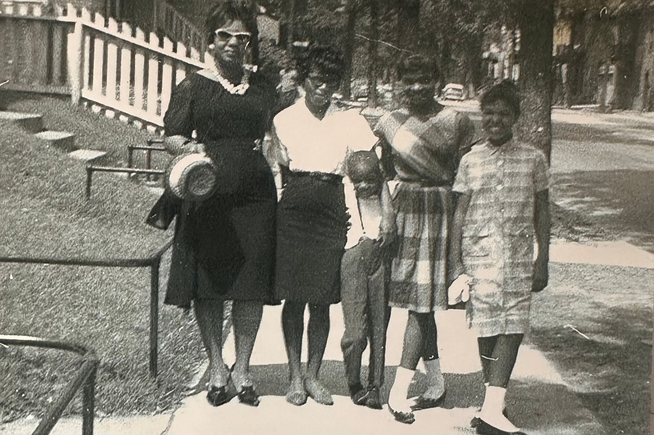 Five women in mid-20th-century dresses stand together on a sidewalk beside a white picket fence, posing for a photo outdoors.