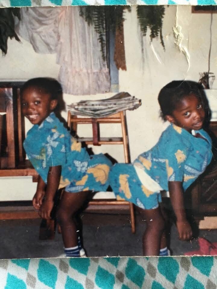 Two young girls in matching blue outfits bend over back-to-back in a playful pose indoors, with a chair and magazines in the background.