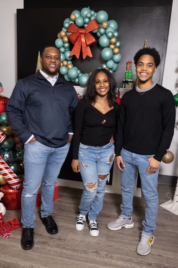 Three smiling people pose for a holiday photo indoors, a man on the left, a woman center, and a young man on the right, in front of a large teal wreath with gold accents and a red bow, Christmas decorations around them.