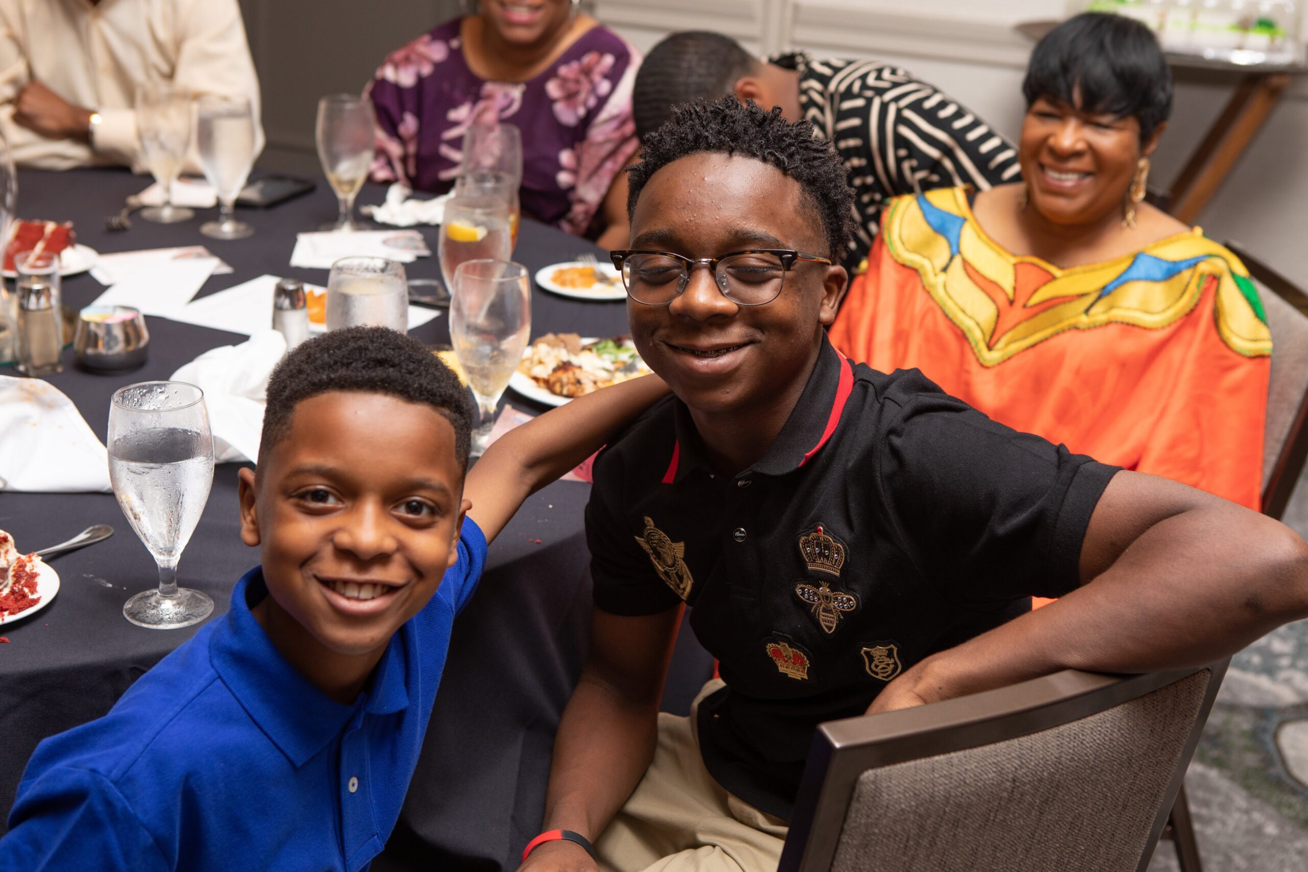 Two smiling boys sit at a dining table with drinks and plates, posing for a photo at a social gathering. In the background, adults chat and laugh, creating a warm party atmosphere.