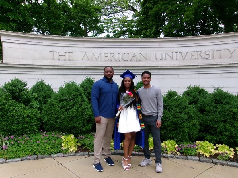 Graduate in blue cap and gown posing with two relatives in front of a curved stone wall that reads 'The American University' while holding flowers