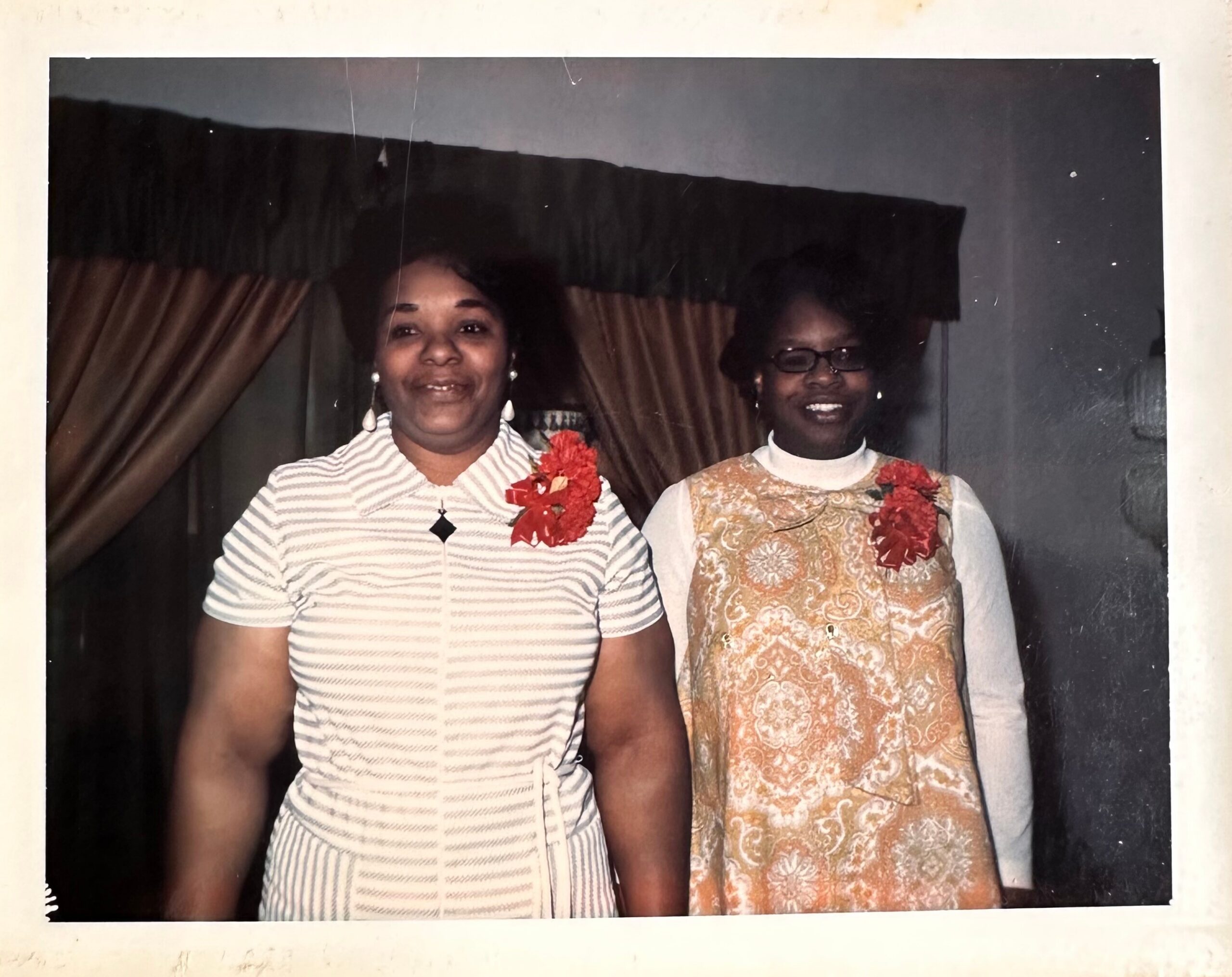 Two women pose together in a framed photo, each wearing festive outfits and red floral accessories, standing in front of brown drapes.