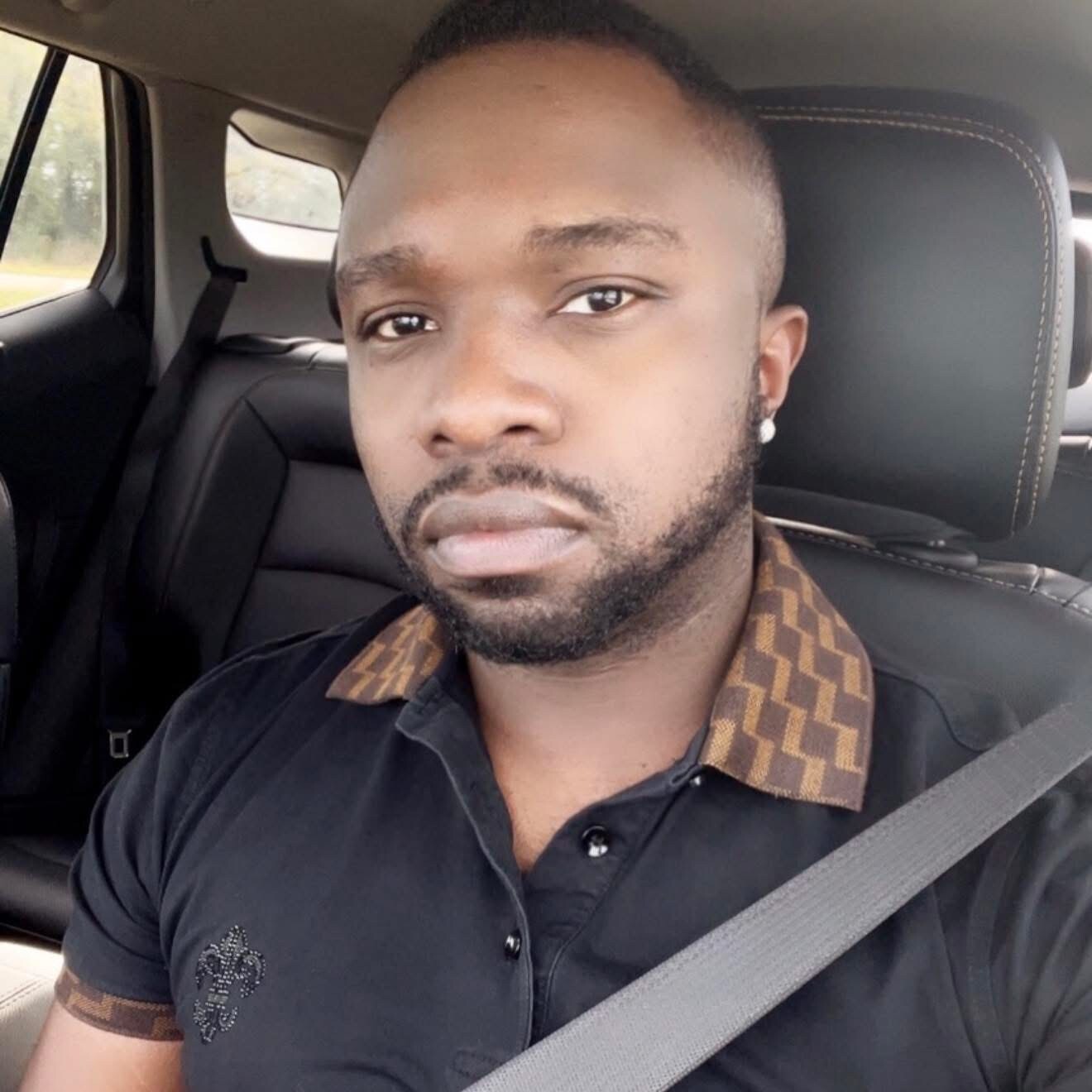 Man in a car wearing a seatbelt, black polo with a brown patterned collar, looking at the camera.