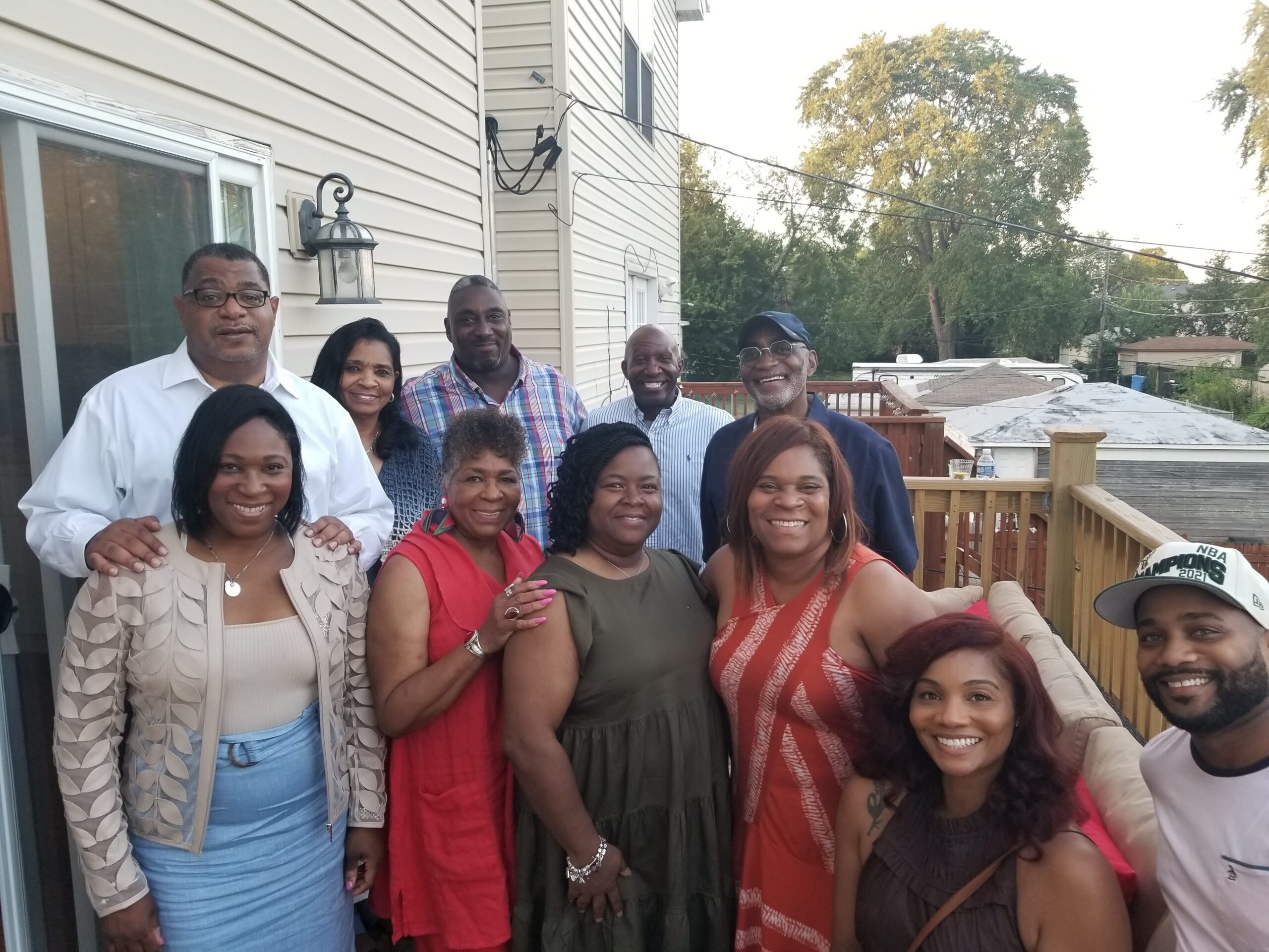 A diverse group of adults posing for a group photo on a wooden deck outside a beige-siding house, smiling at the camera.