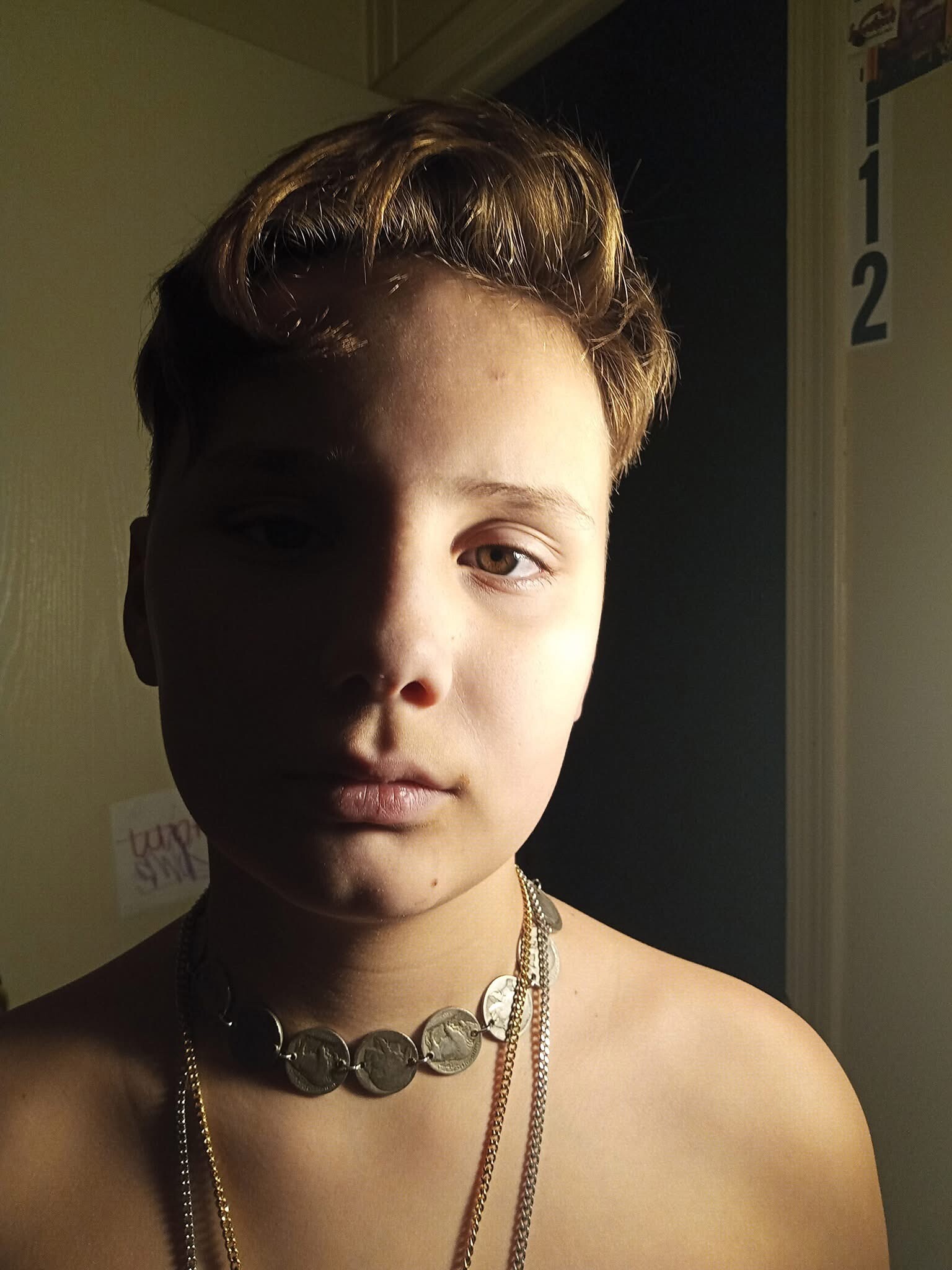 Young person with short, light brown hair wearing layered gold chains, bare shoulders, in a dimly lit room with a doorway marked '2' in the background.