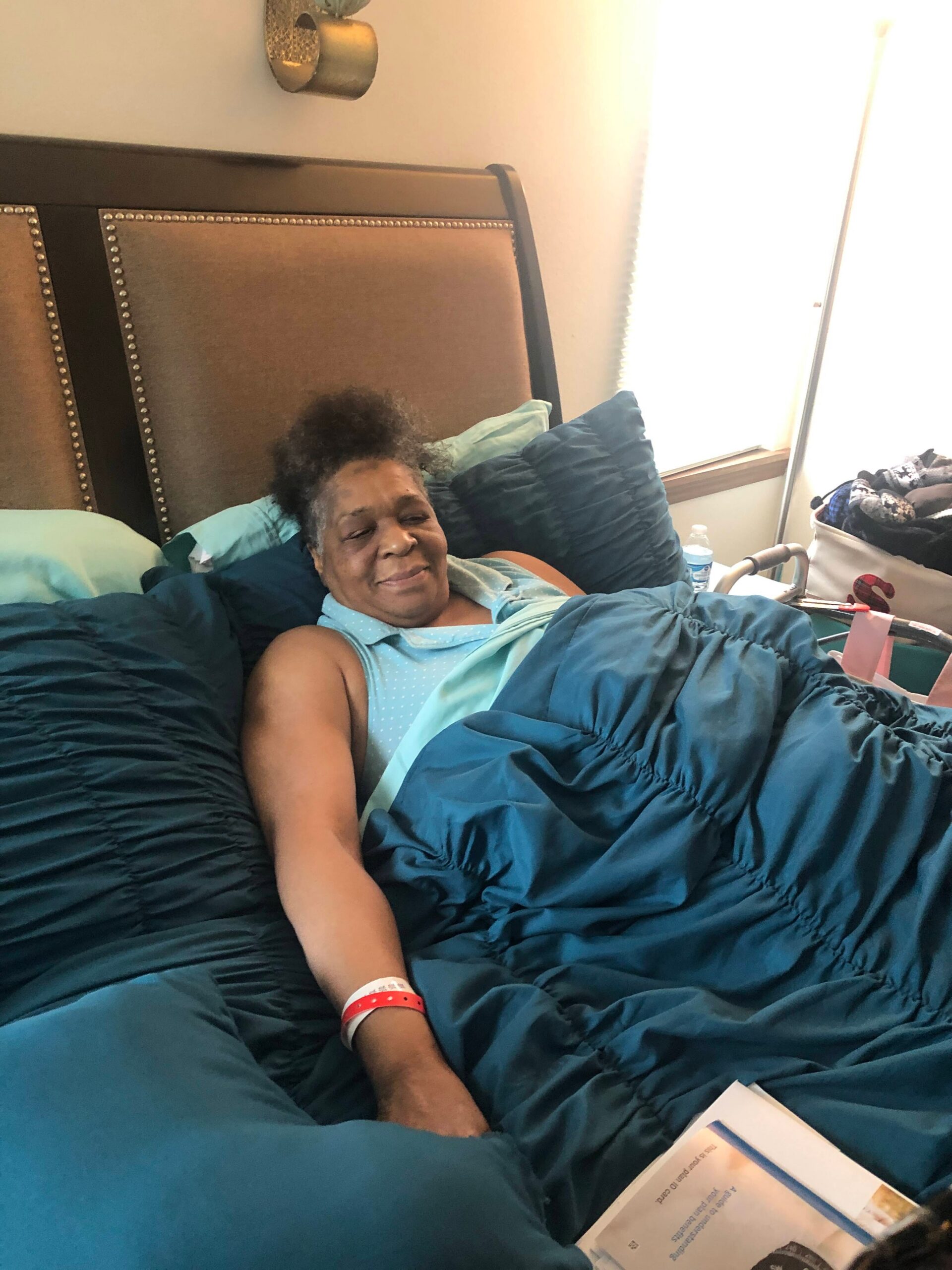 Older woman resting in a hospital bed, smiling, tucked in a blue-green blanket with teal pillows nearby.