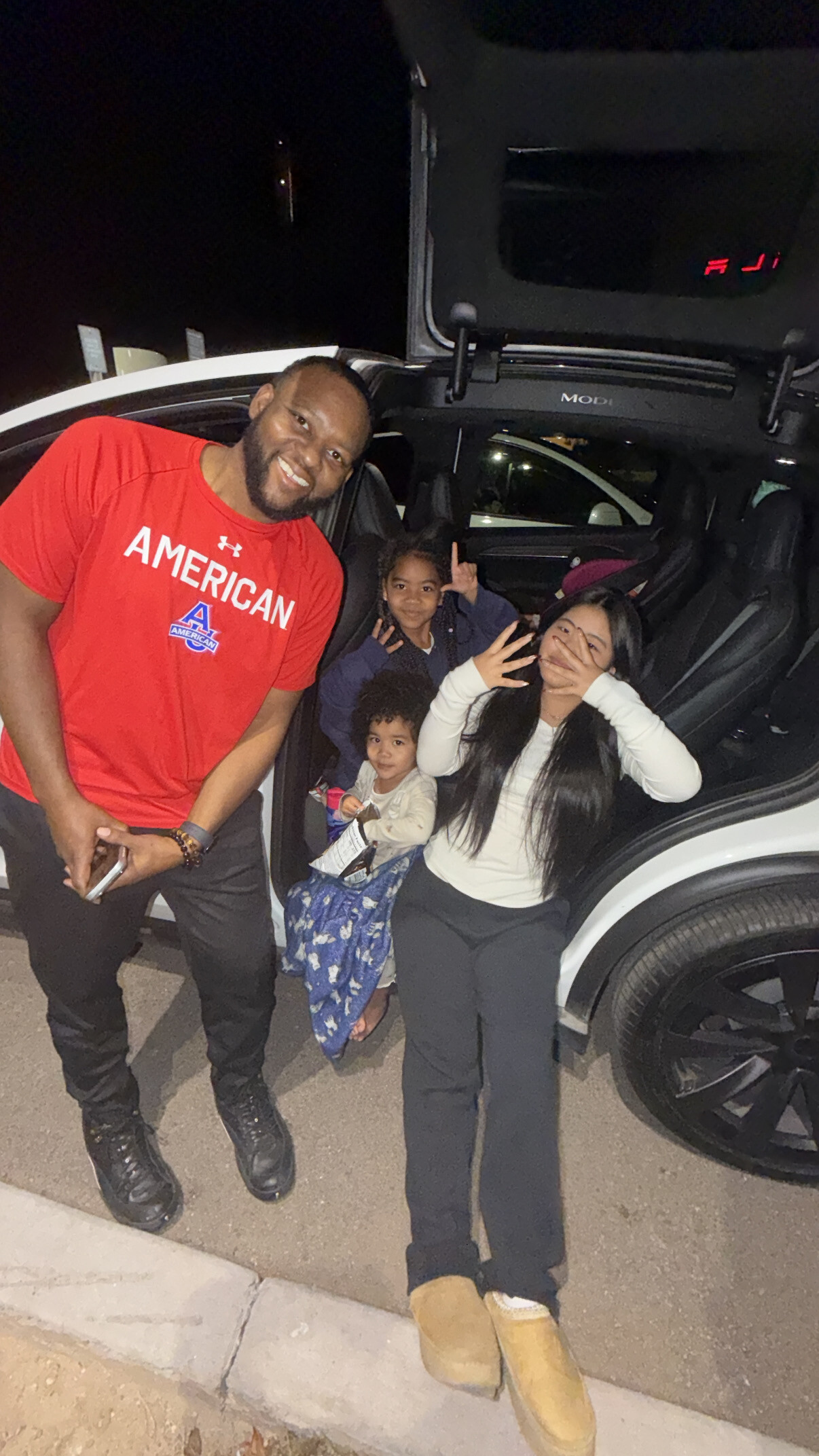 A smiling man in a red American shirt standing next to a white SUV with its open trunk, accompanied by a woman and two children posing at night.