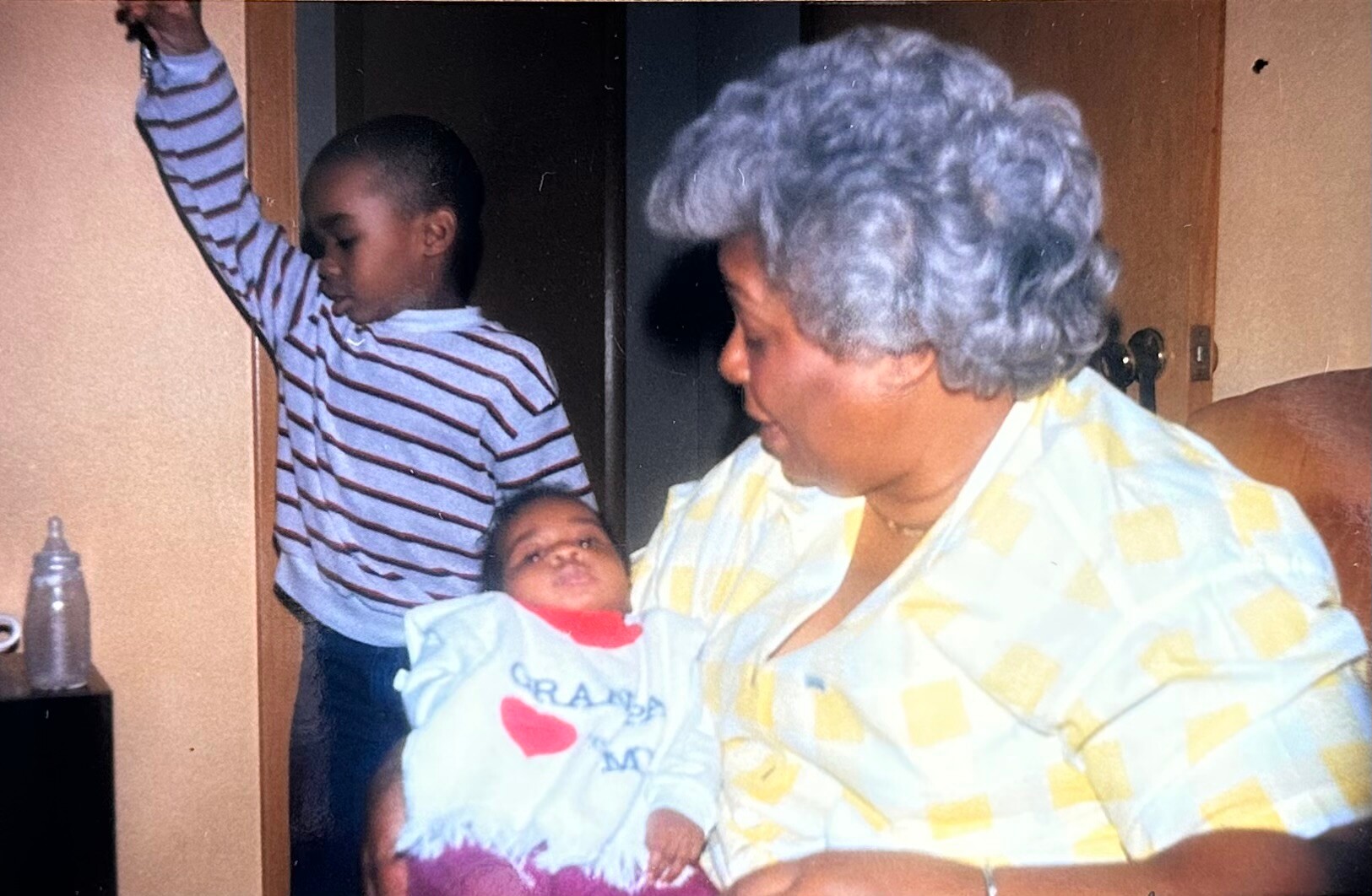 Grandmother with gray hair holding a newborn, while a boy in a striped shirt stands near a doorway in a home.