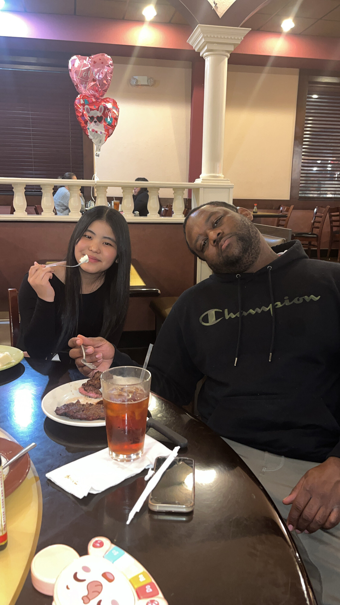 Young woman and man dine at a restaurant; pink heart-shaped Valentine balloons float behind them.