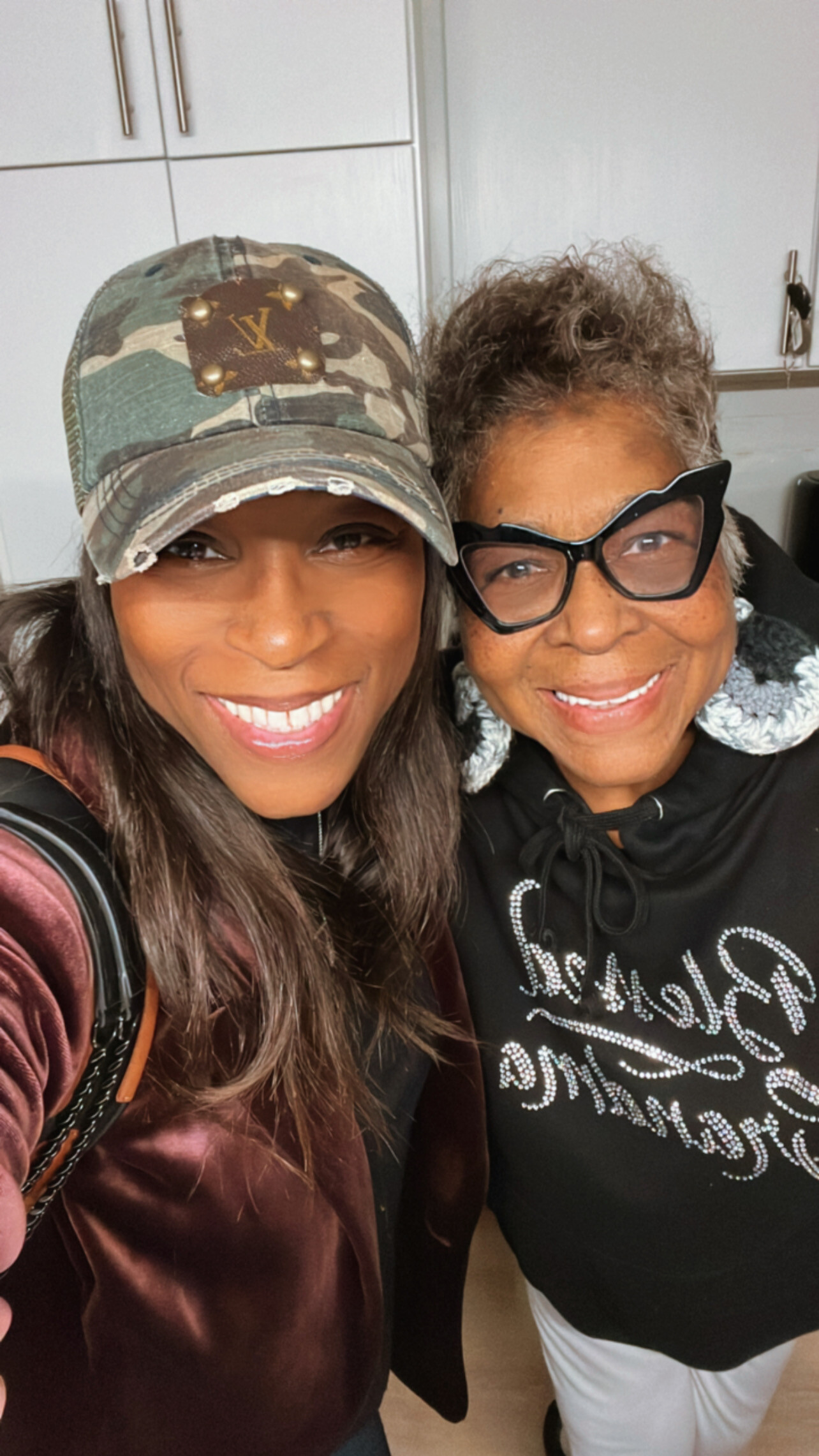 Two women smiling for a selfie in a kitchen; left woman wears a camo cap and burgundy velvet jacket, right woman wears large black glasses and a black top with white writing.