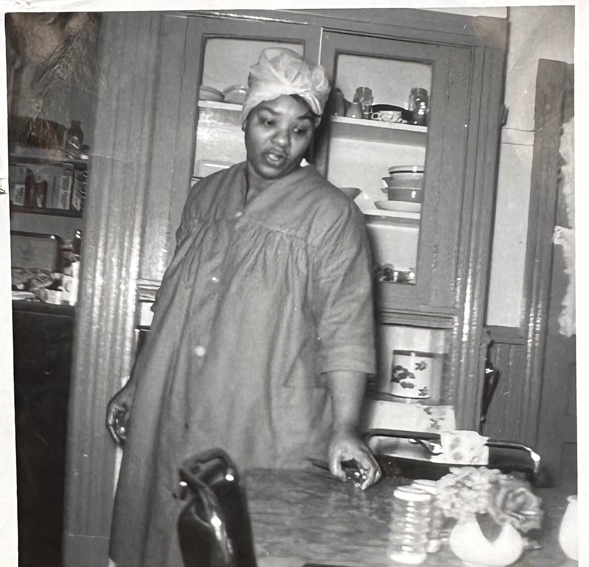 Black-and-white photo of a woman in a loose hospital-style gown and head wrap standing beside a kitchen counter, looking to the side, with a cabinet of dishes visible behind her.