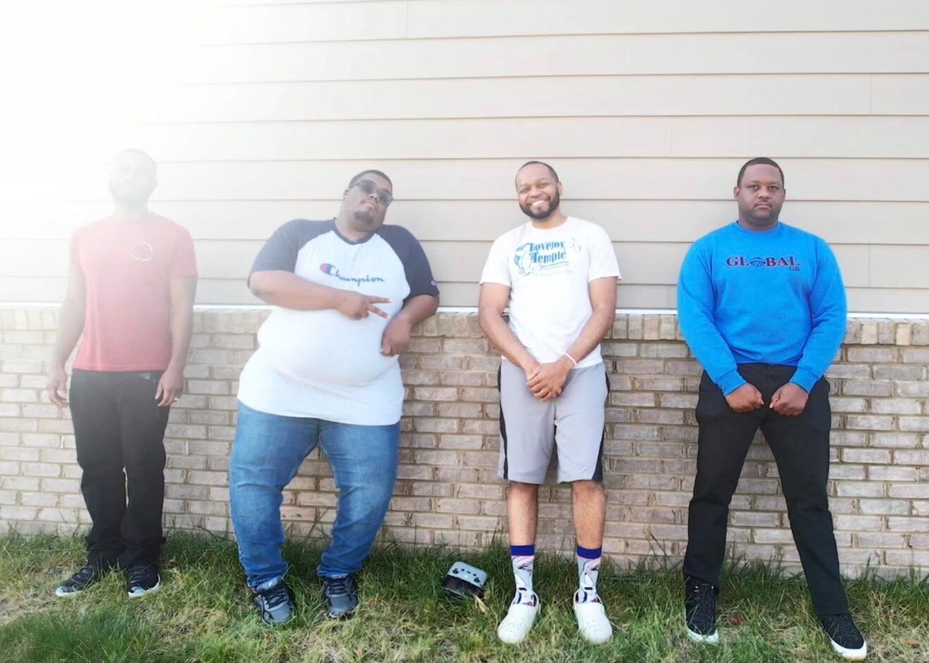 Four men pose against a beige brick wall: one in a red shirt, another overweight man in a light gray Champion tee, a third man in a white graphic shirt with shorts, and a fourth man in a blue sweatshirt.