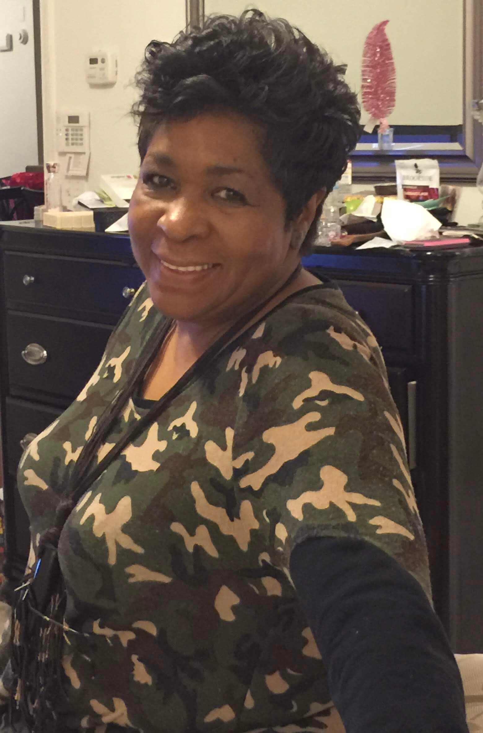Smiling woman with short black hair wearing a camouflage top in an office setting.