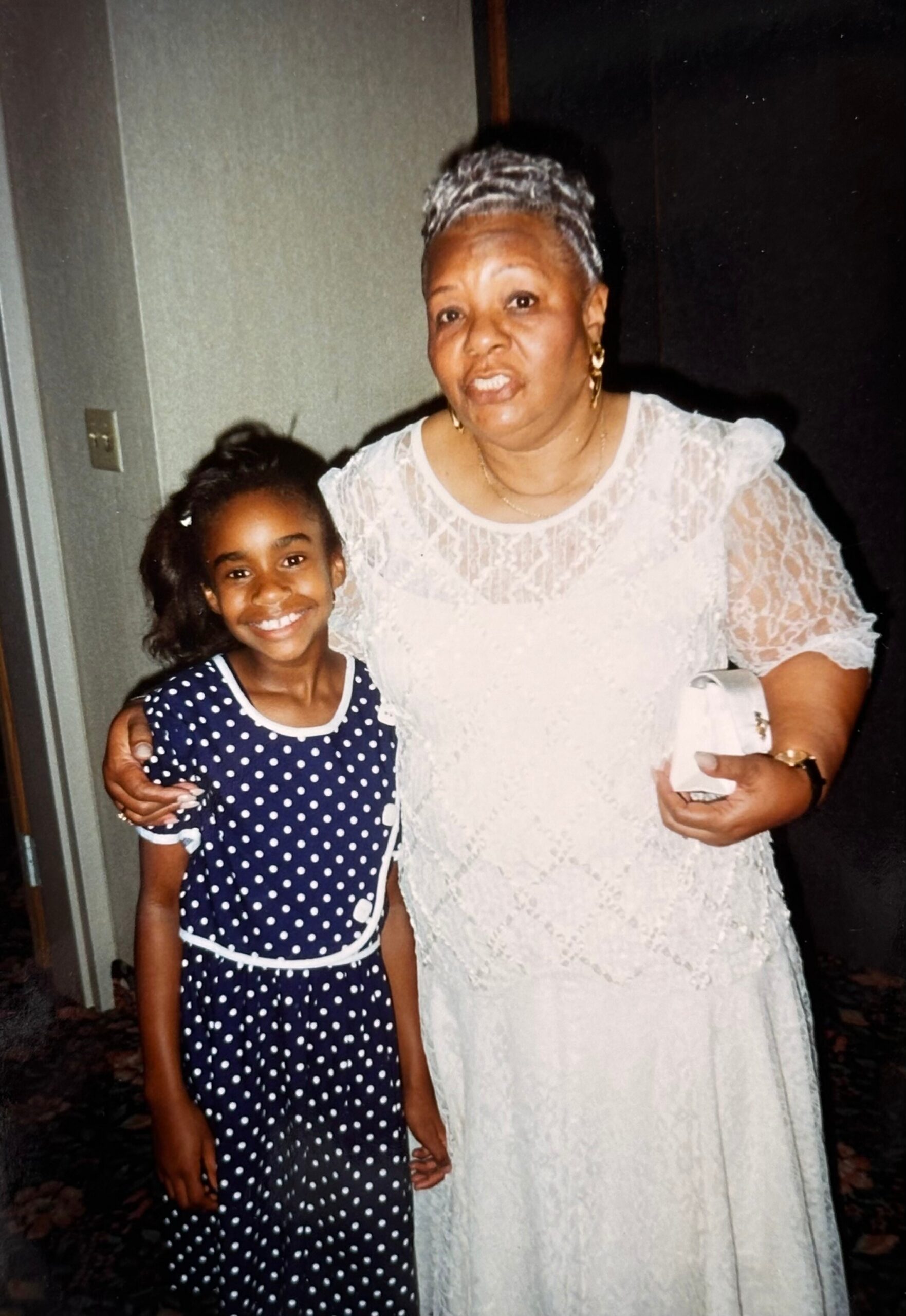 Grandmother and young granddaughter pose for a photo indoors, smiling together; the grandmother wears a white lace dress and gold jewelry, the girl wears a navy polka-dot dress.