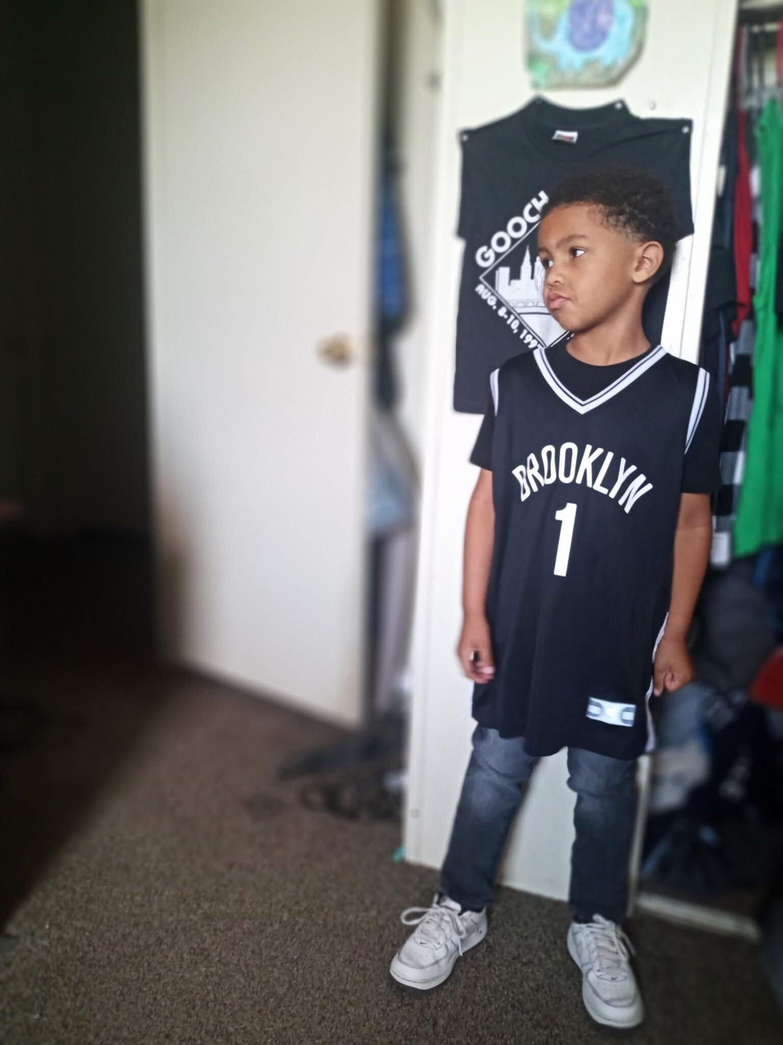 Boy wearing a black Brooklyn basketball jersey and white sneakers stands in a doorway indoors, with a closet in the background.