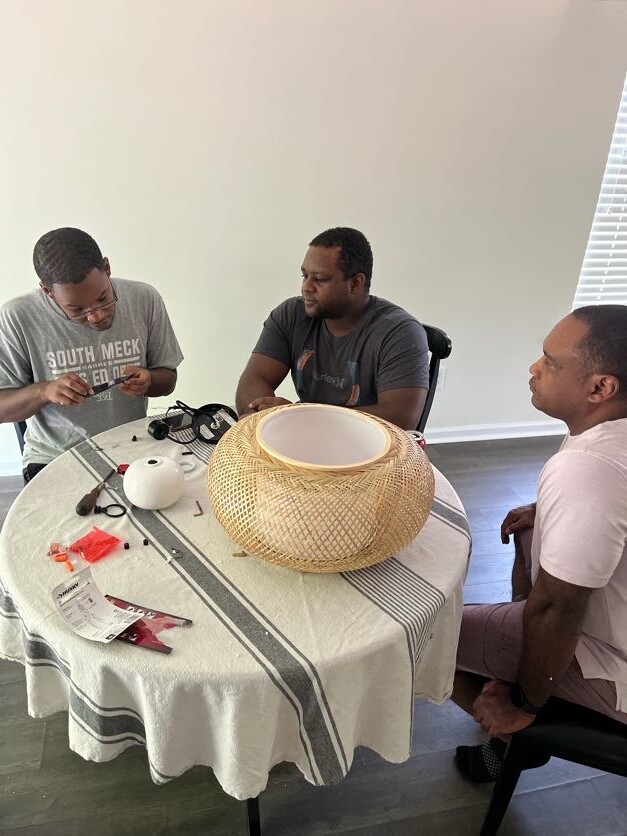 Three men sit around a table assembling a large woven wicker basket, with tools and materials on the table in a casual workshop setting.