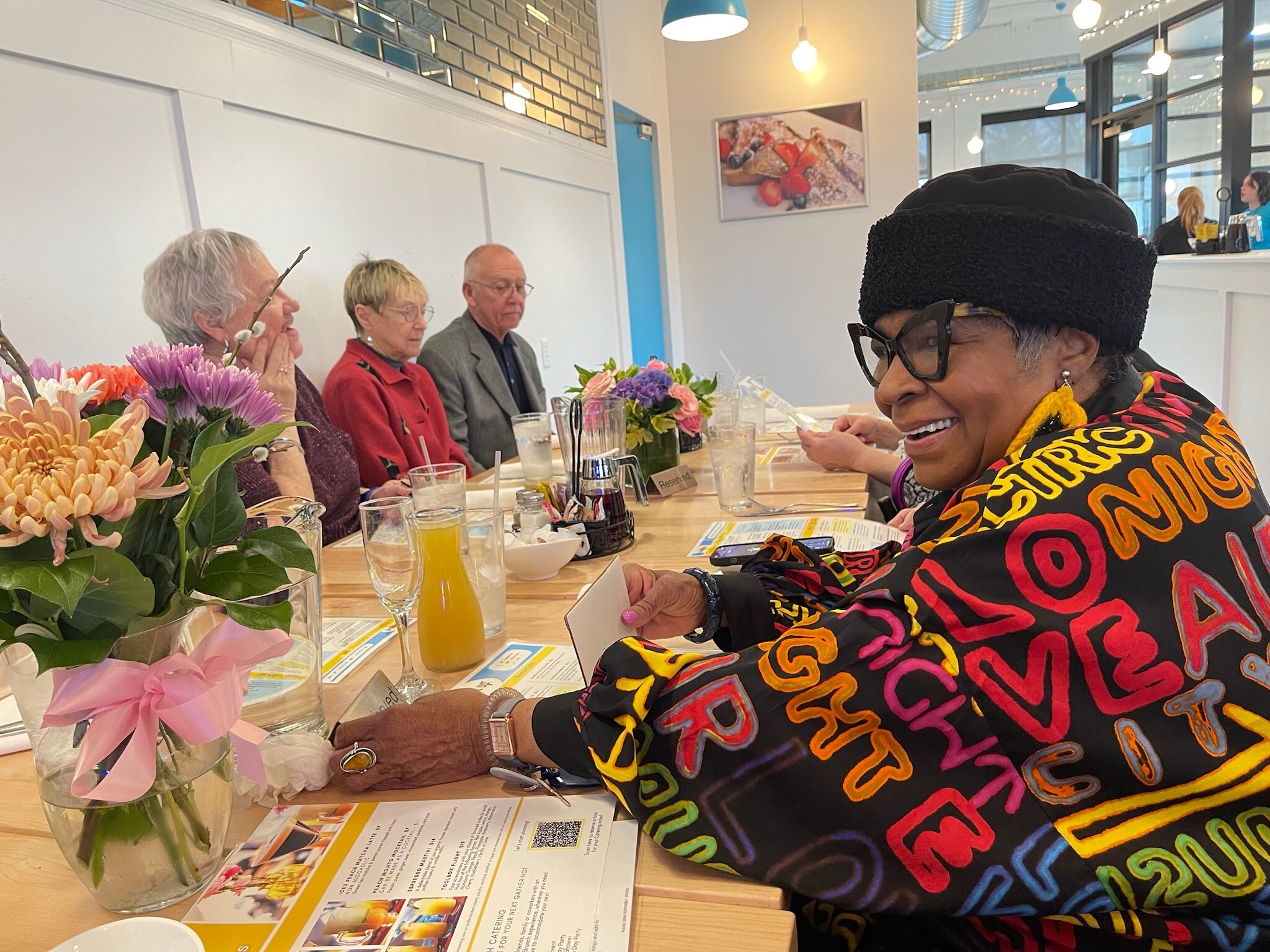 People dining at a long table with flowers, talking and smiling in a bright cafe setting.