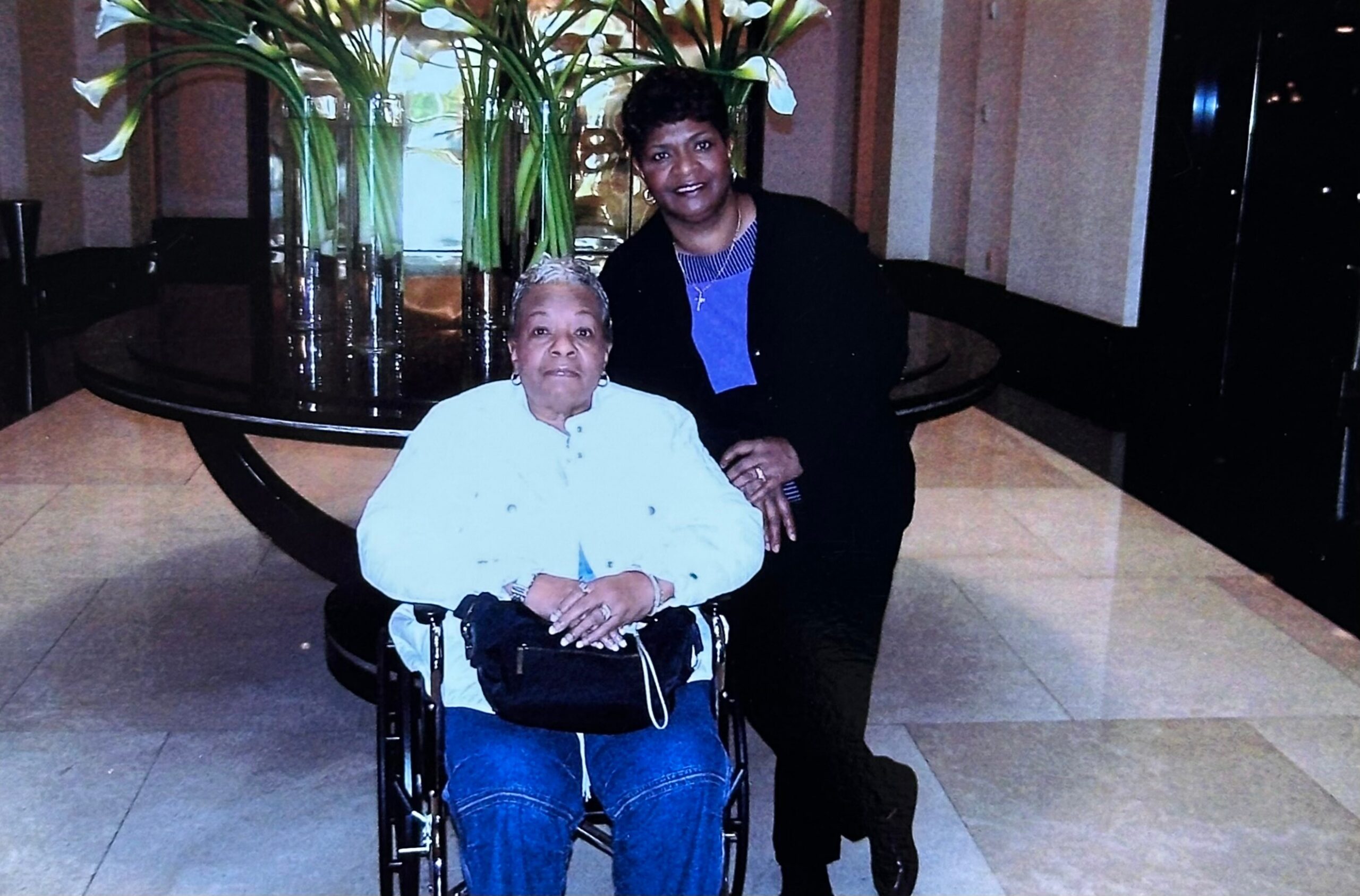 Two women pose together in a lobby; one seated in a wheelchair wearing a white top and blue jeans, the other standing beside her in a black jacket and blue top, large floral arrangement behind them