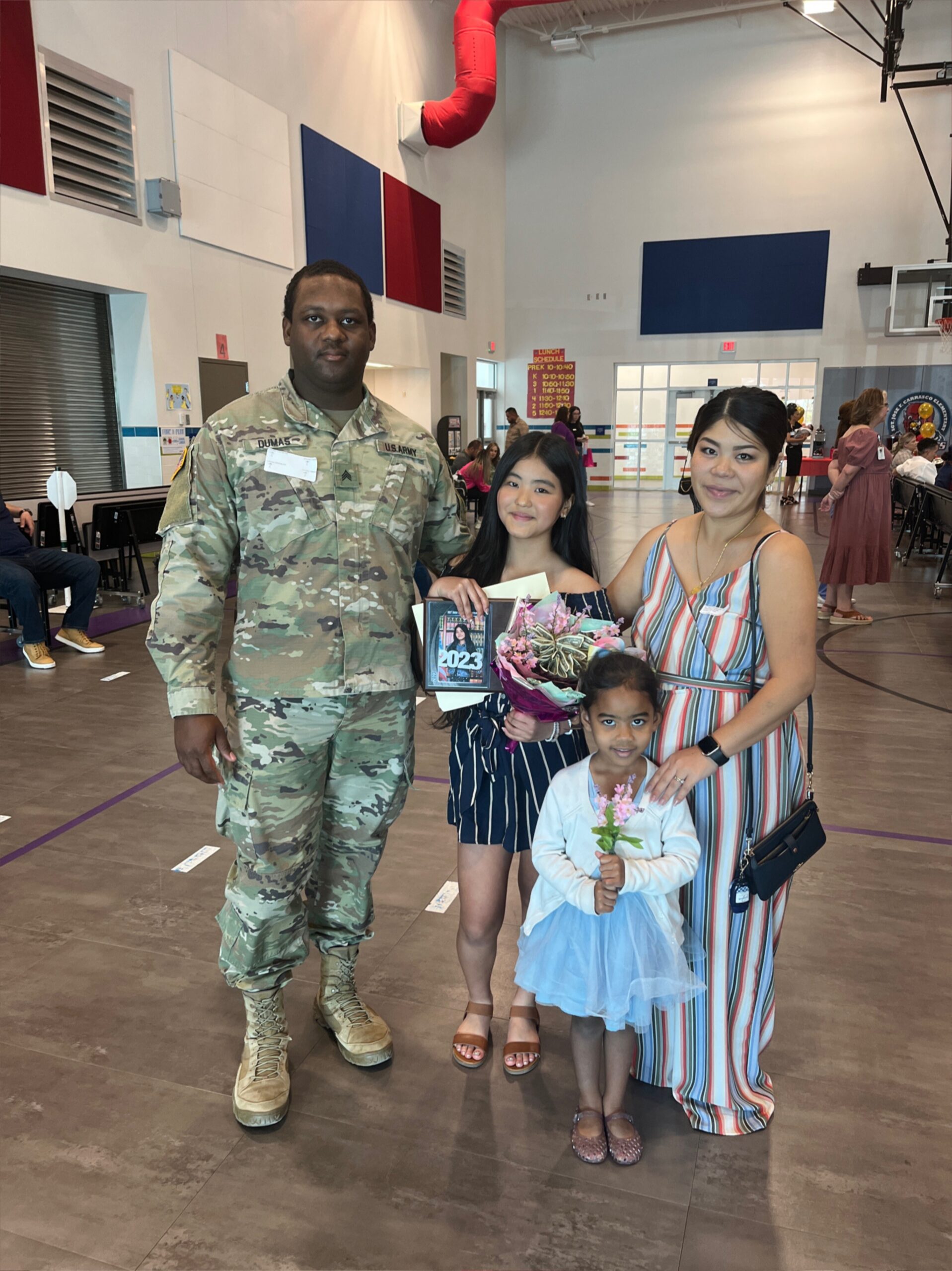 Army soldier in camouflage uniform poses with a smiling mother and two daughters at a school gym, holding flowers and a yearbook/photos.