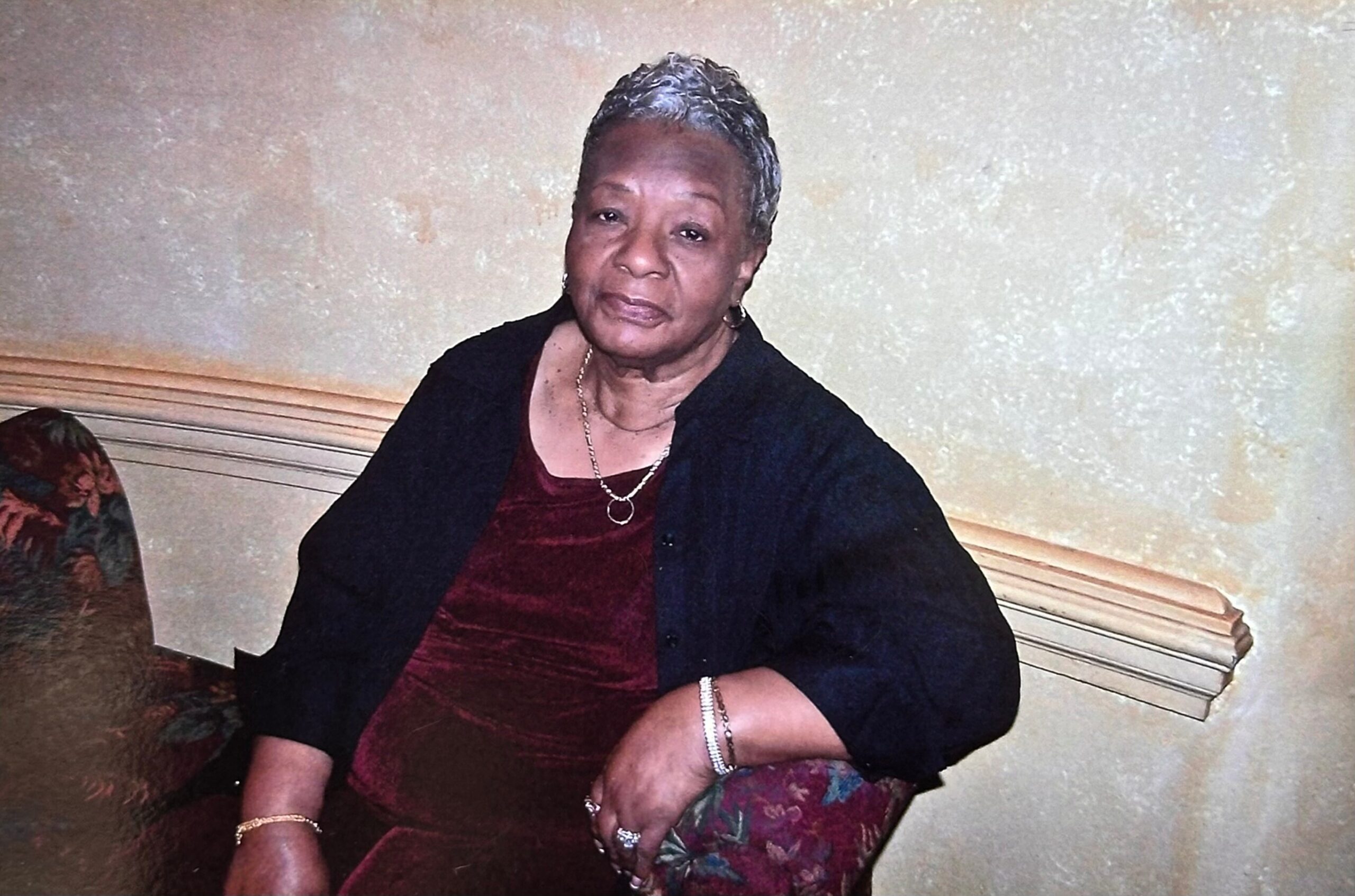 Elderly woman with short gray hair wearing a burgundy dress and black cardigan, seated on a wooden bench indoors.