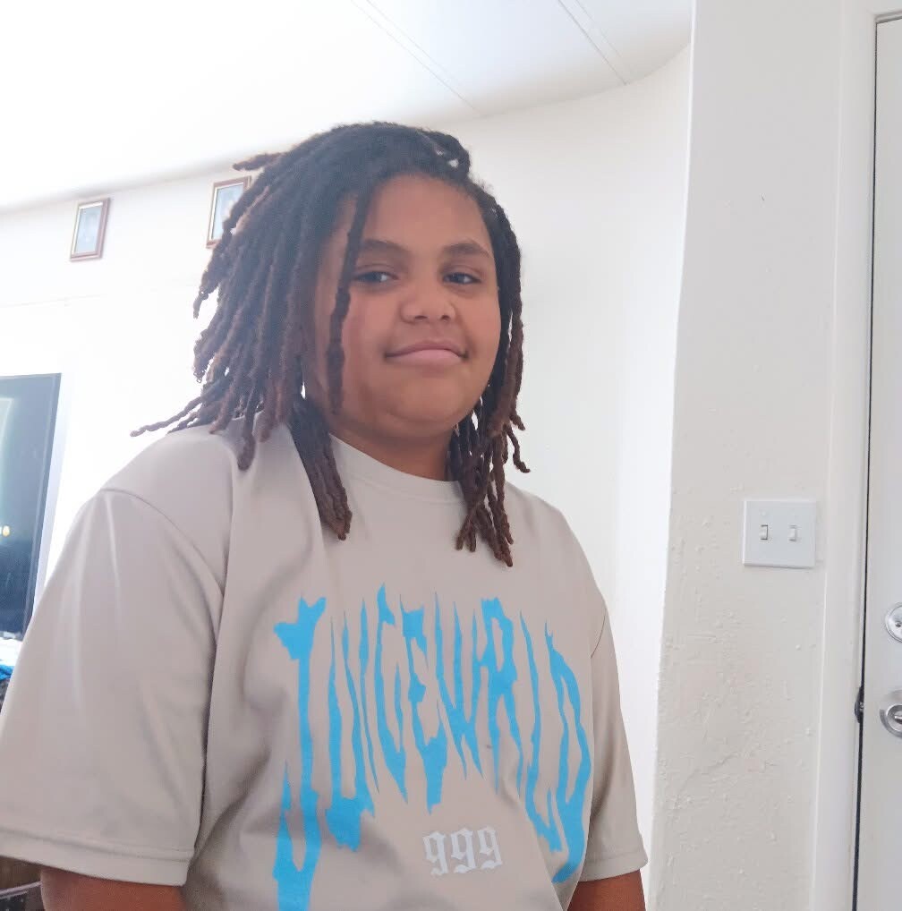 Person with shoulder-length dreadlocks wearing a light gray graphic T-shirt, standing indoors and smiling slightly.