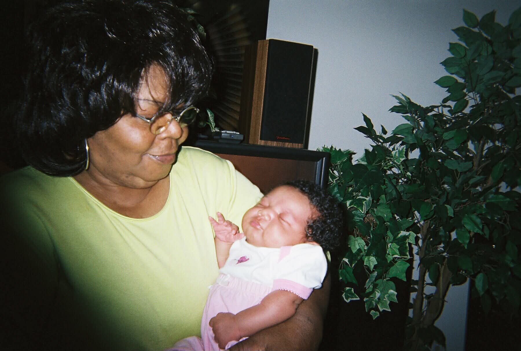 Smiling grandmother in a lime green top holds a newborn baby girl indoors, with a leafy plant in the background.