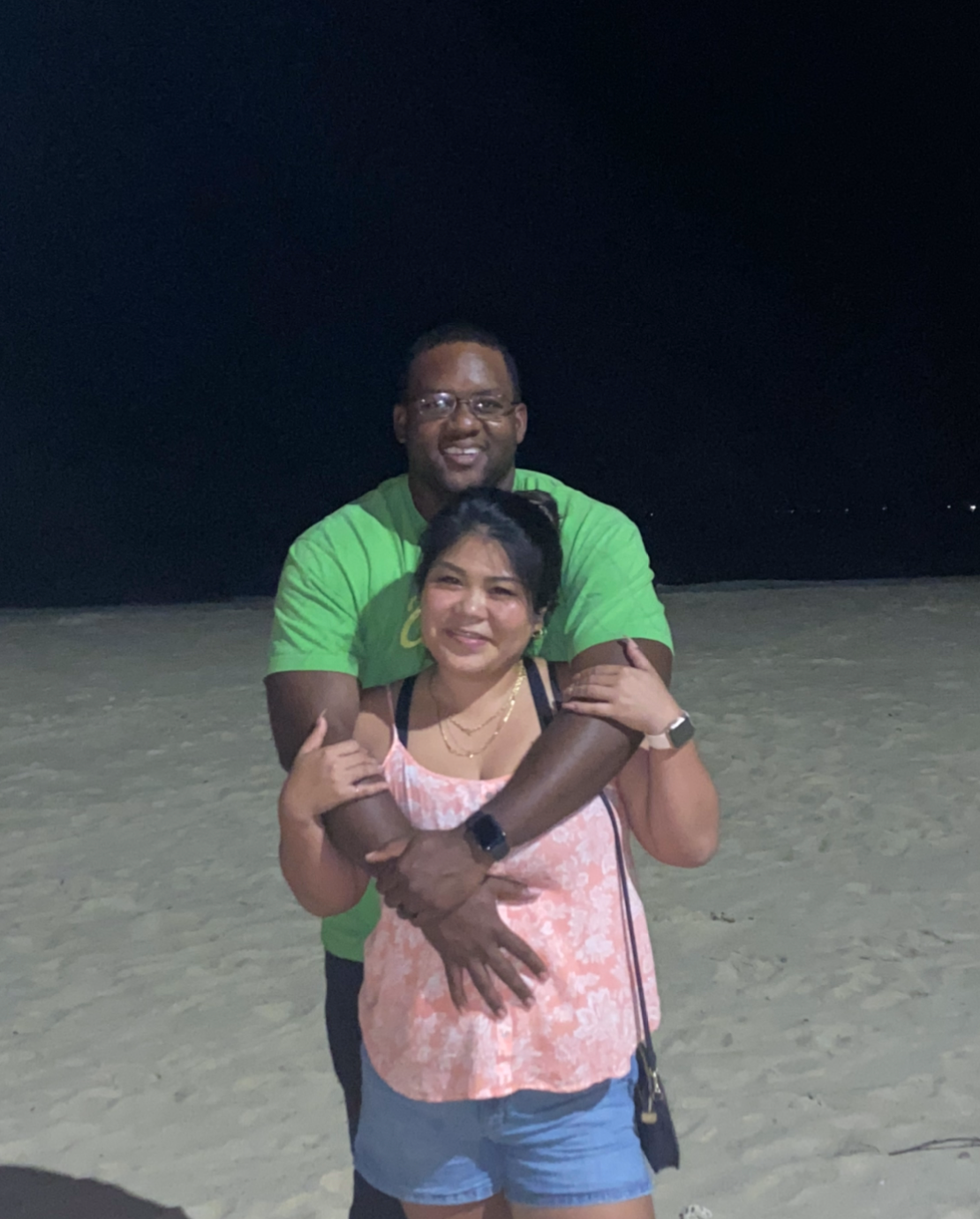 Smiling couple on a sandy beach at night; man in a bright green shirt hugs a woman in a peach top from behind.