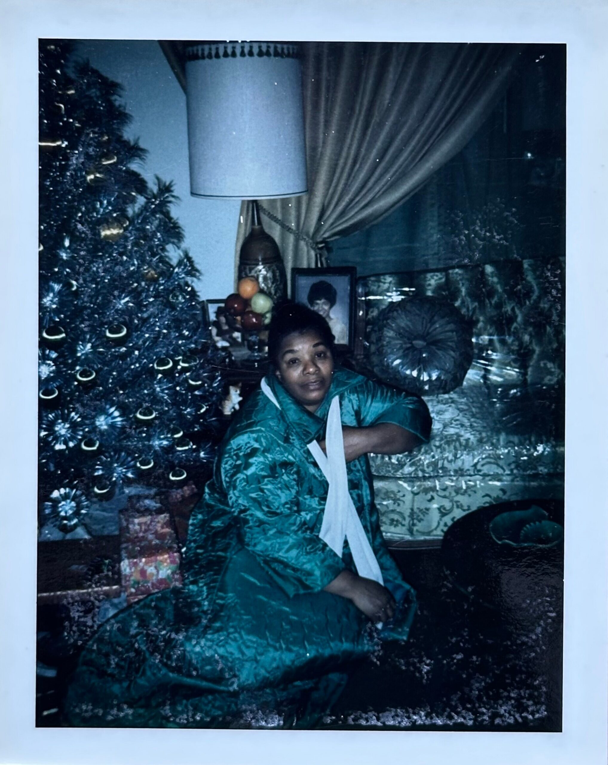 A woman kneels beside a decorated Christmas tree indoors, with presents beneath and framed photos on the table in the background.