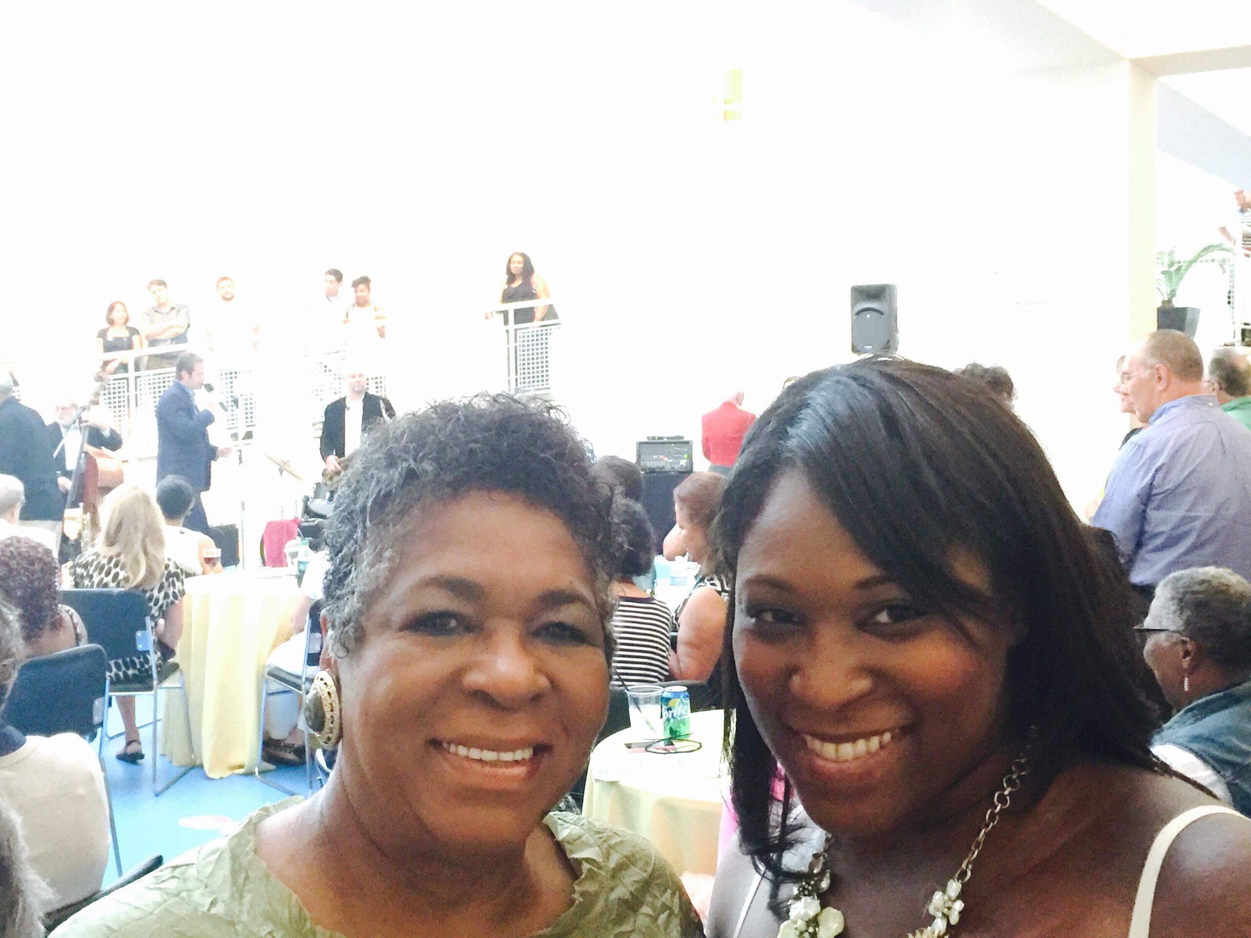 Two women smiling for a selfie in a crowded indoor event, with people standing and sitting in the background and a stage area on the left.