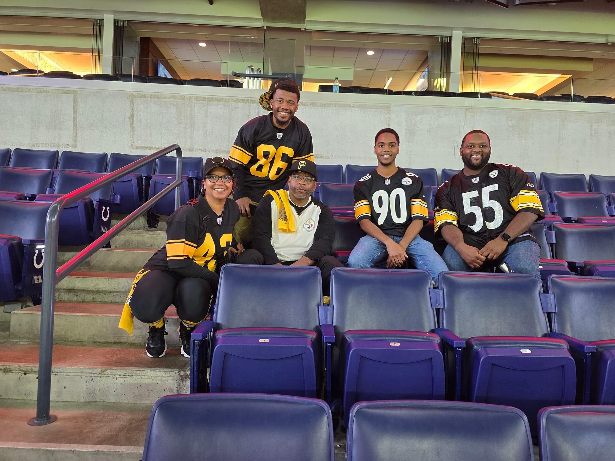 Group of friends and family wearing Pittsburgh Steelers jerseys posing and smiling in stadium seating.