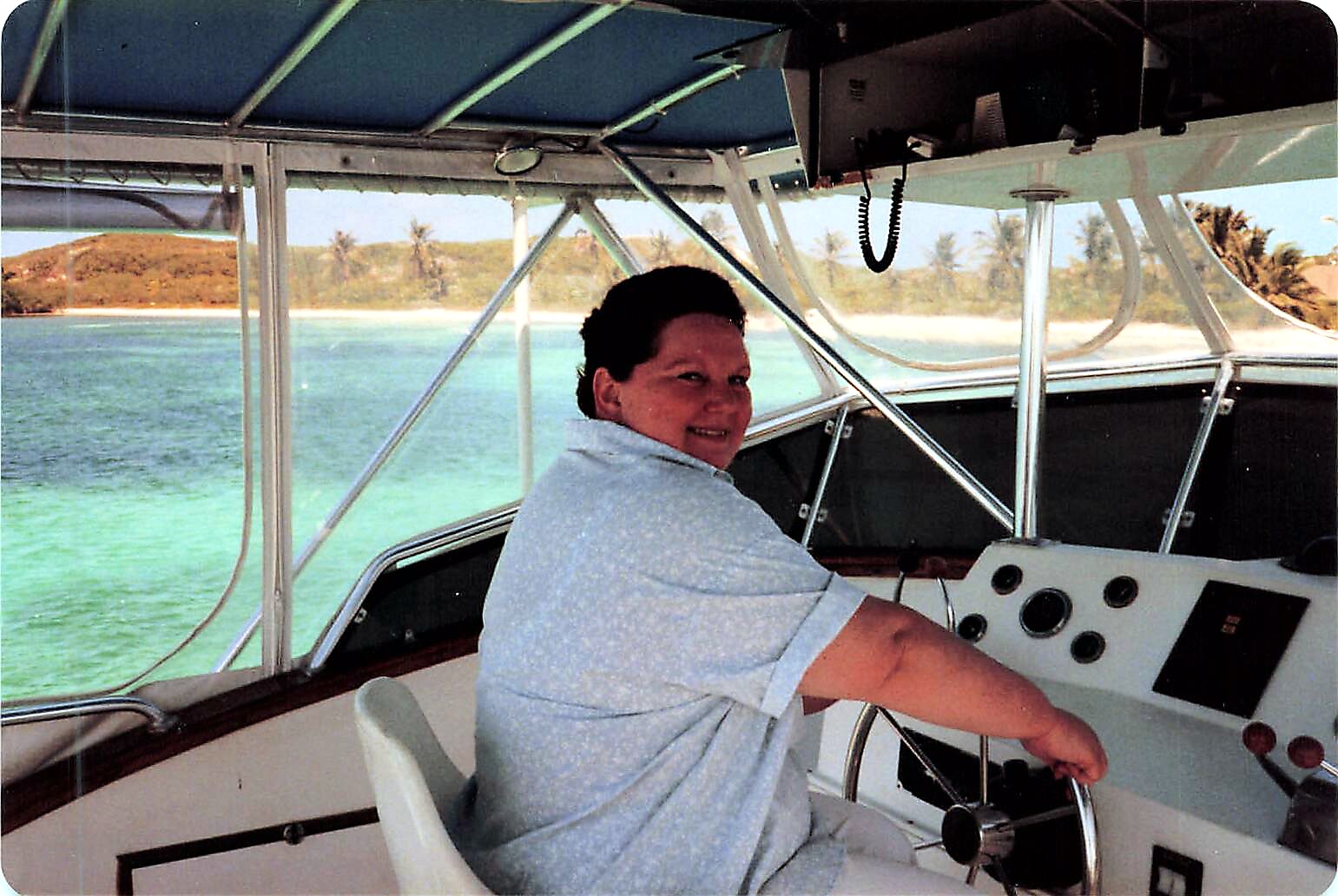 Smiling woman steering a boat at the helm with turquoise water and palm trees in the background.